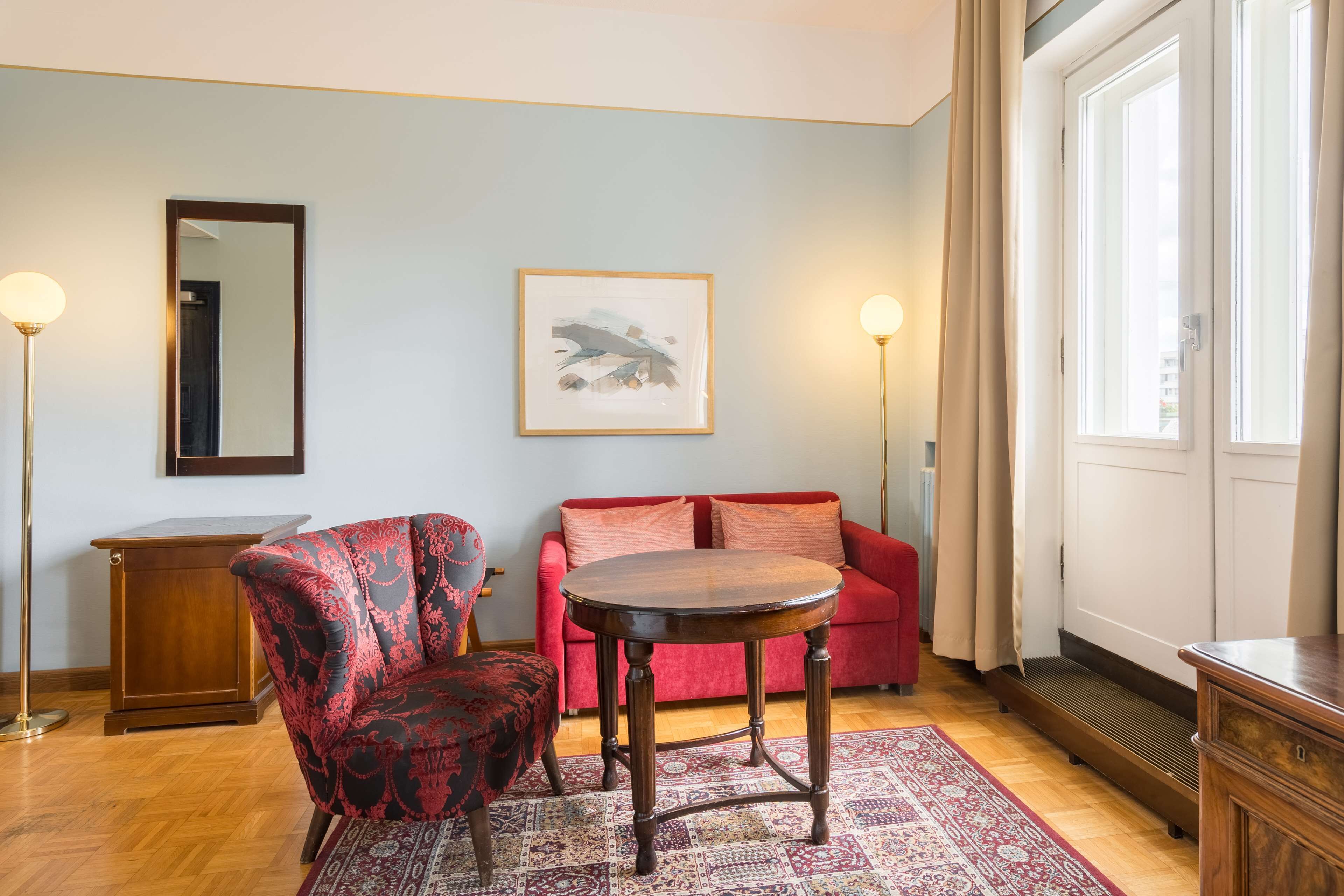 Una habitación con un sofá rojo, una mesa redonda de madera, un sillón estampado, un espejo y un cuadro abstracto enmarcado en la pared.