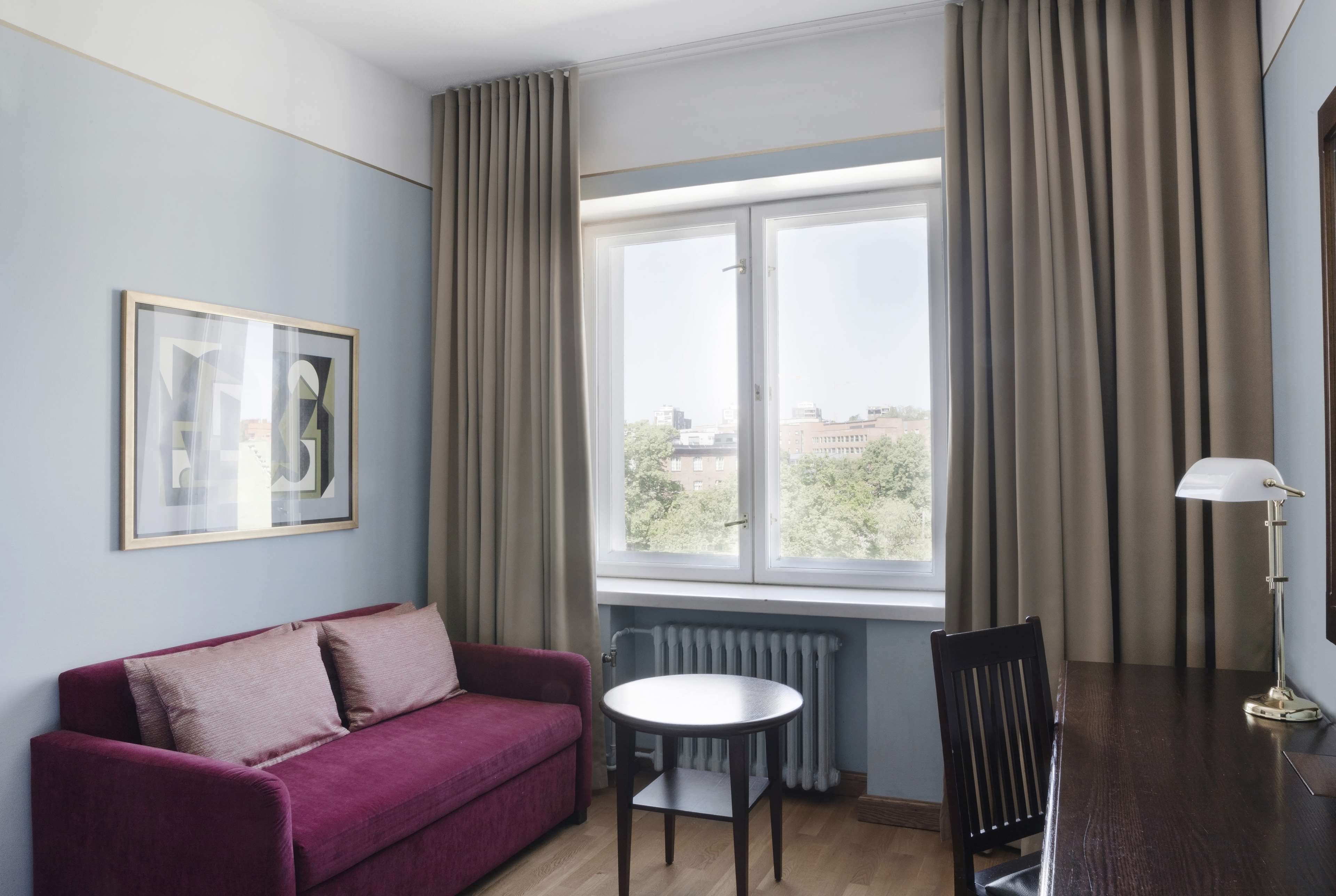 A room with a red sofa, a small round table, a chair, a radiator, and a window with curtains.