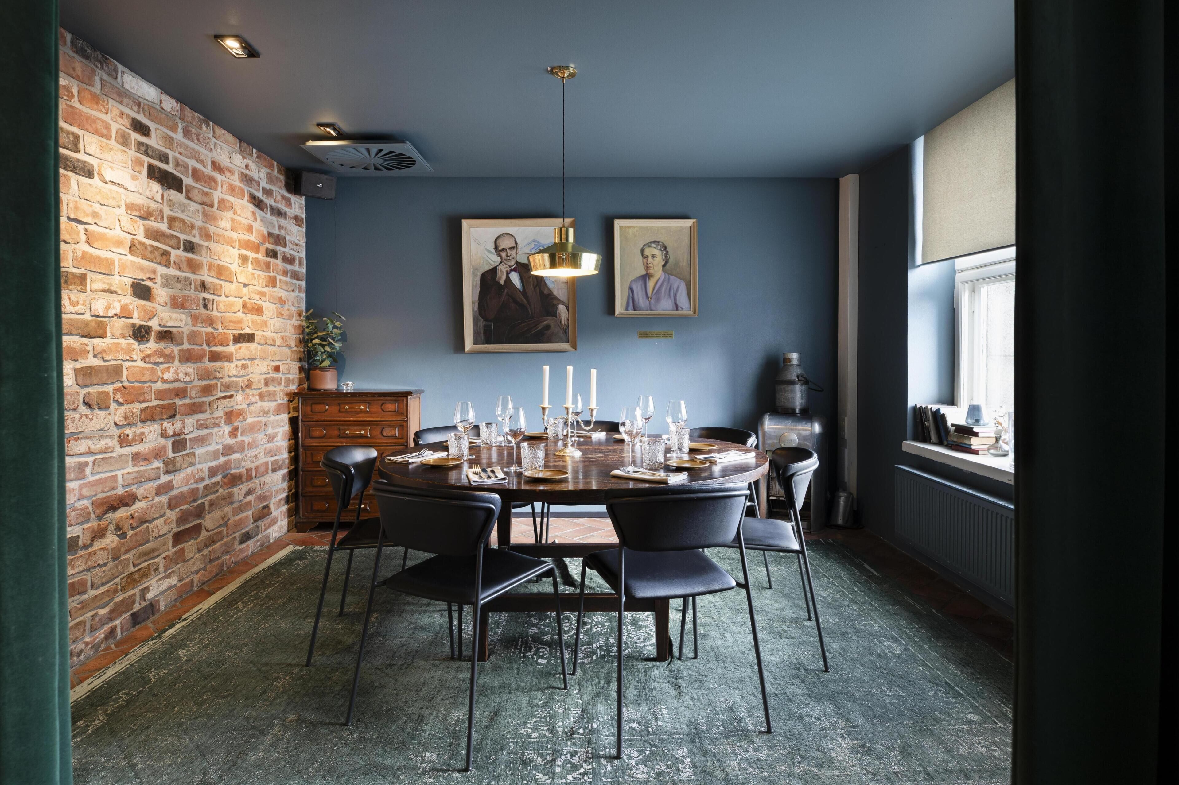 A dining area with a brick wall, a round table set for six, and two framed portraits on the wall.