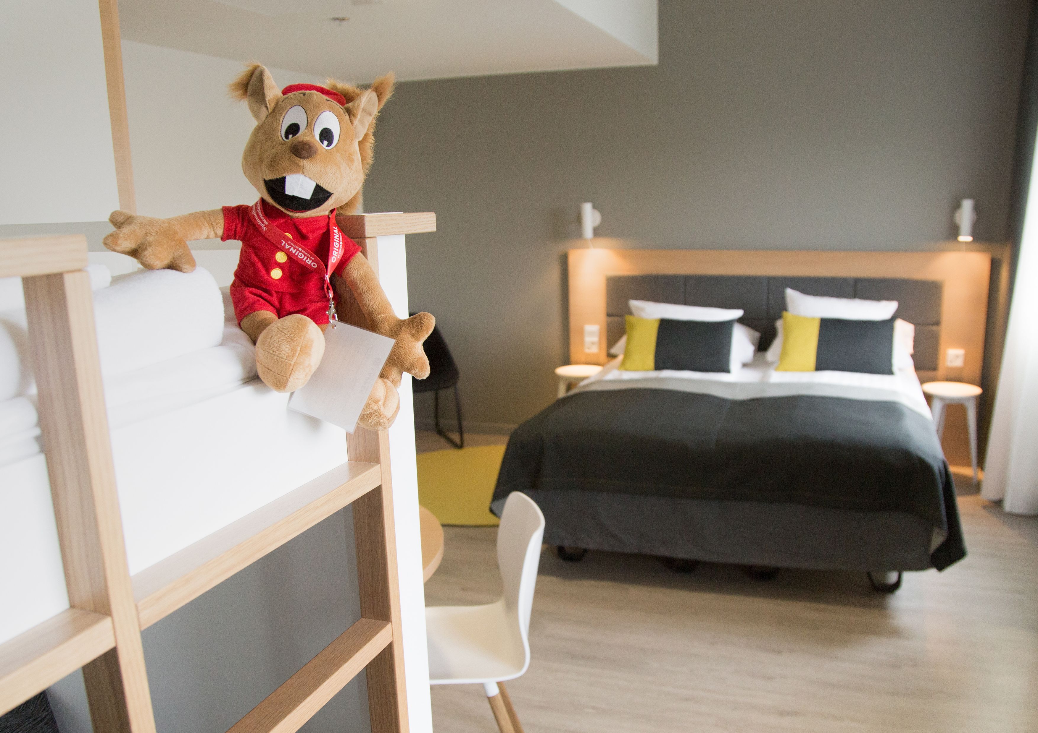 A hotel room with a bed, a desk with a teddy bear, and a chair.