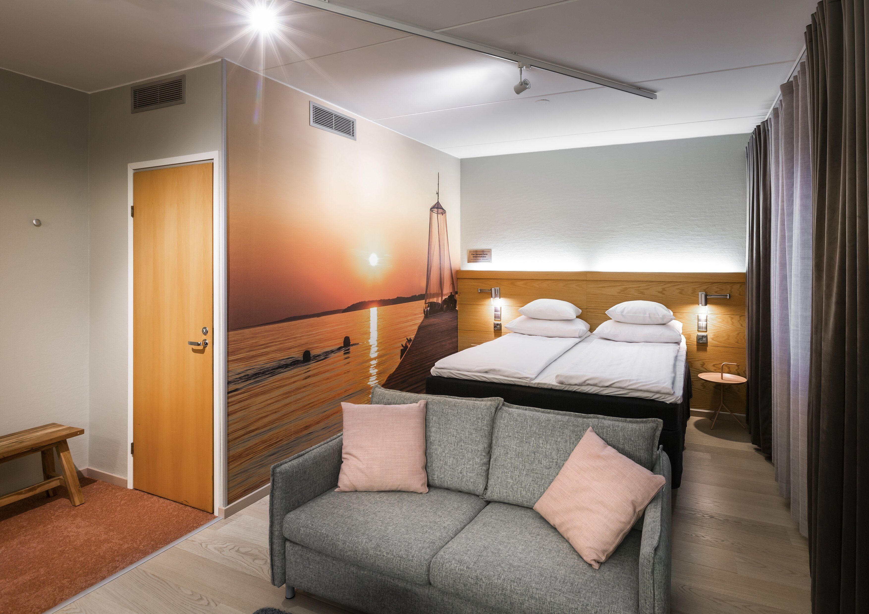 A hotel room with a bed, a sofa, a wall mural, and a small table.