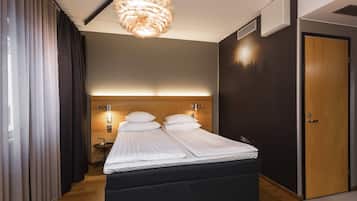 A modern hotel room with a large bed, a wooden headboard, a wall-mounted light, and a door.