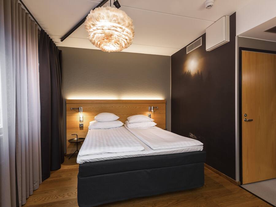 A modern hotel room with a large bed, a wooden headboard, a wall-mounted light, and a door.