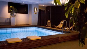 A swimming pool with wooden deck and lounge chairs, surrounded by walls with a dark curtain and a mounted TV. - Original Sokos Hotel Lappee (Lappeenranta)