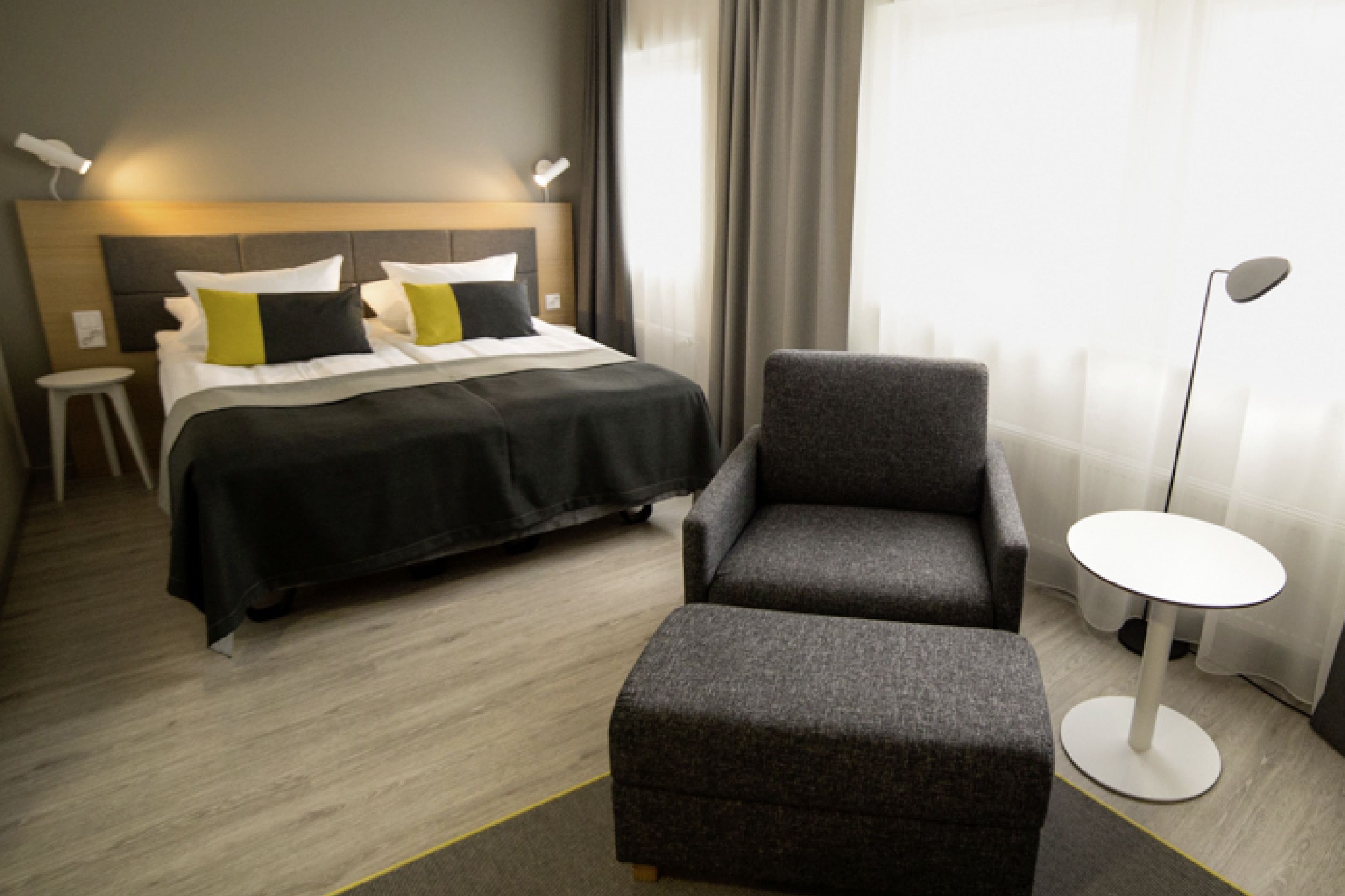A hotel room with a bed, a grey armchair, a small white table, and a floor lamp.