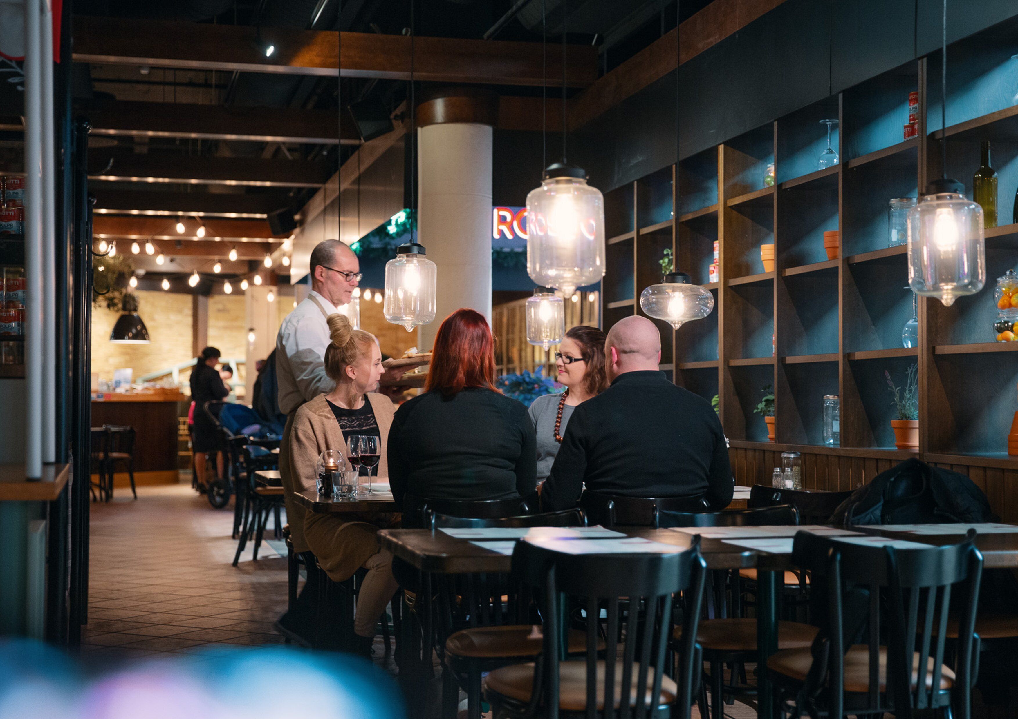 A restaurant interior with tables, chairs, hanging light fixtures, and shelves with glassware.