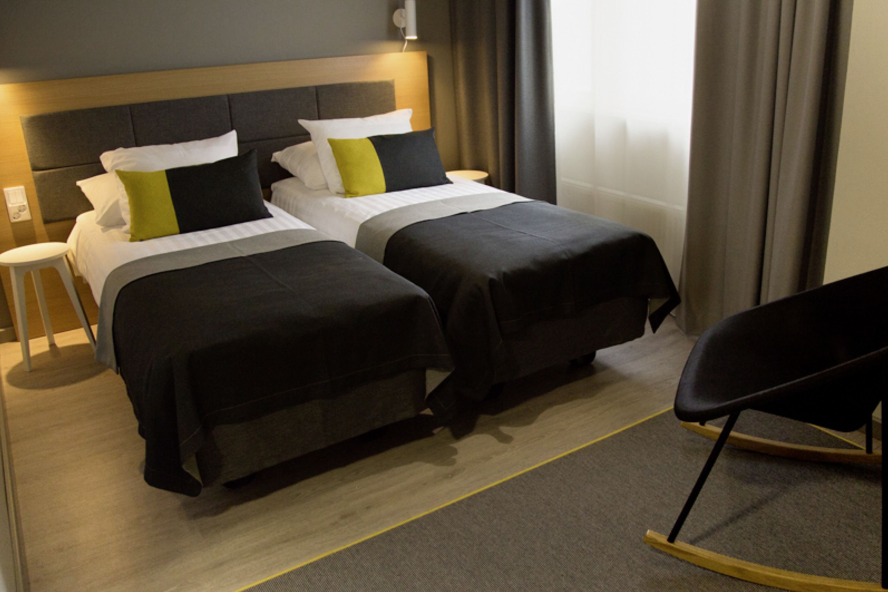 Habitació d'hotel amb dos llits, una meseta de nit, una cadira i un capçal amb il·luminació integrada.