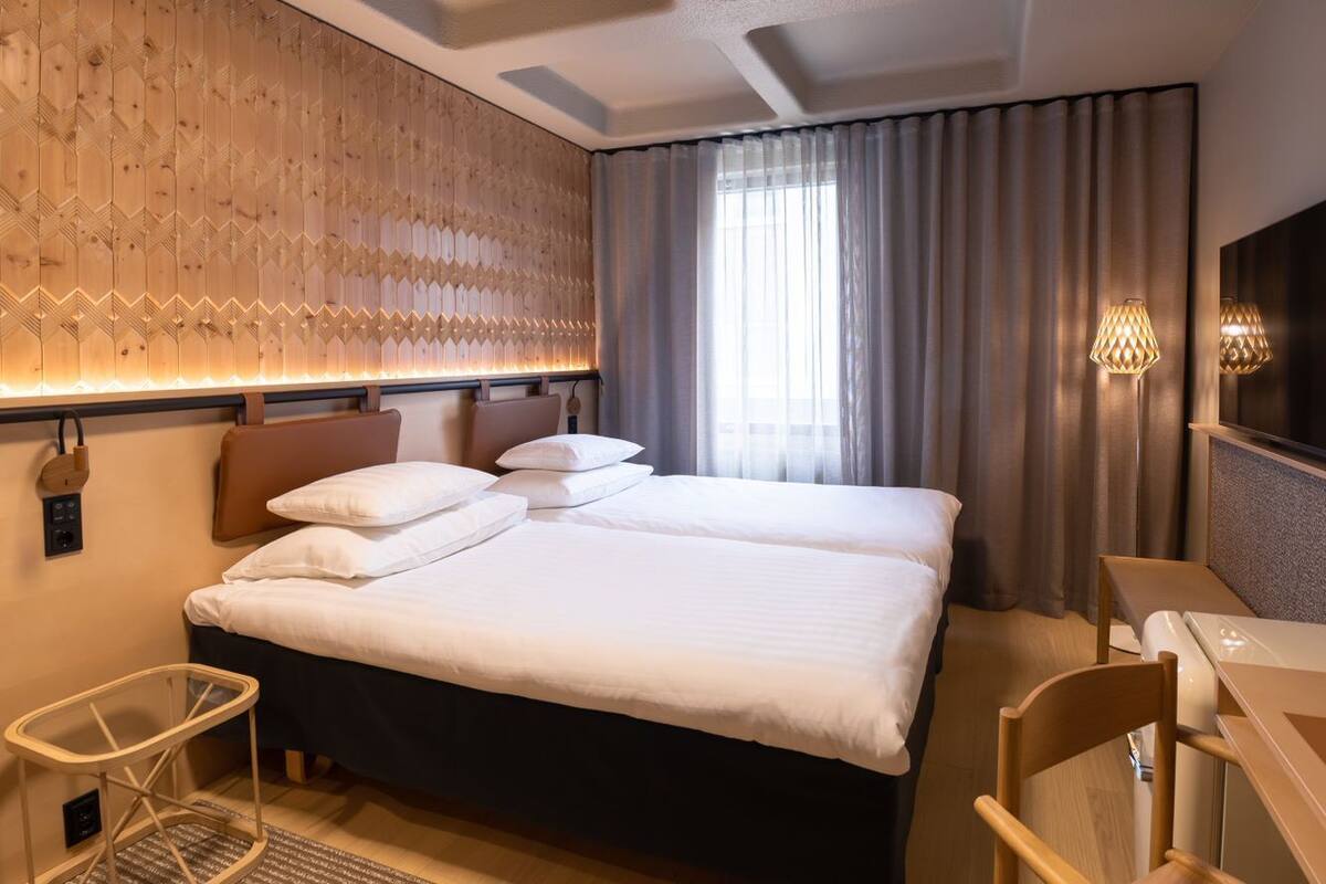 Quarto de hotel com uma cama grande, uma mesa, uma cadeira, uma TV e uma parede com padrão geométrico.