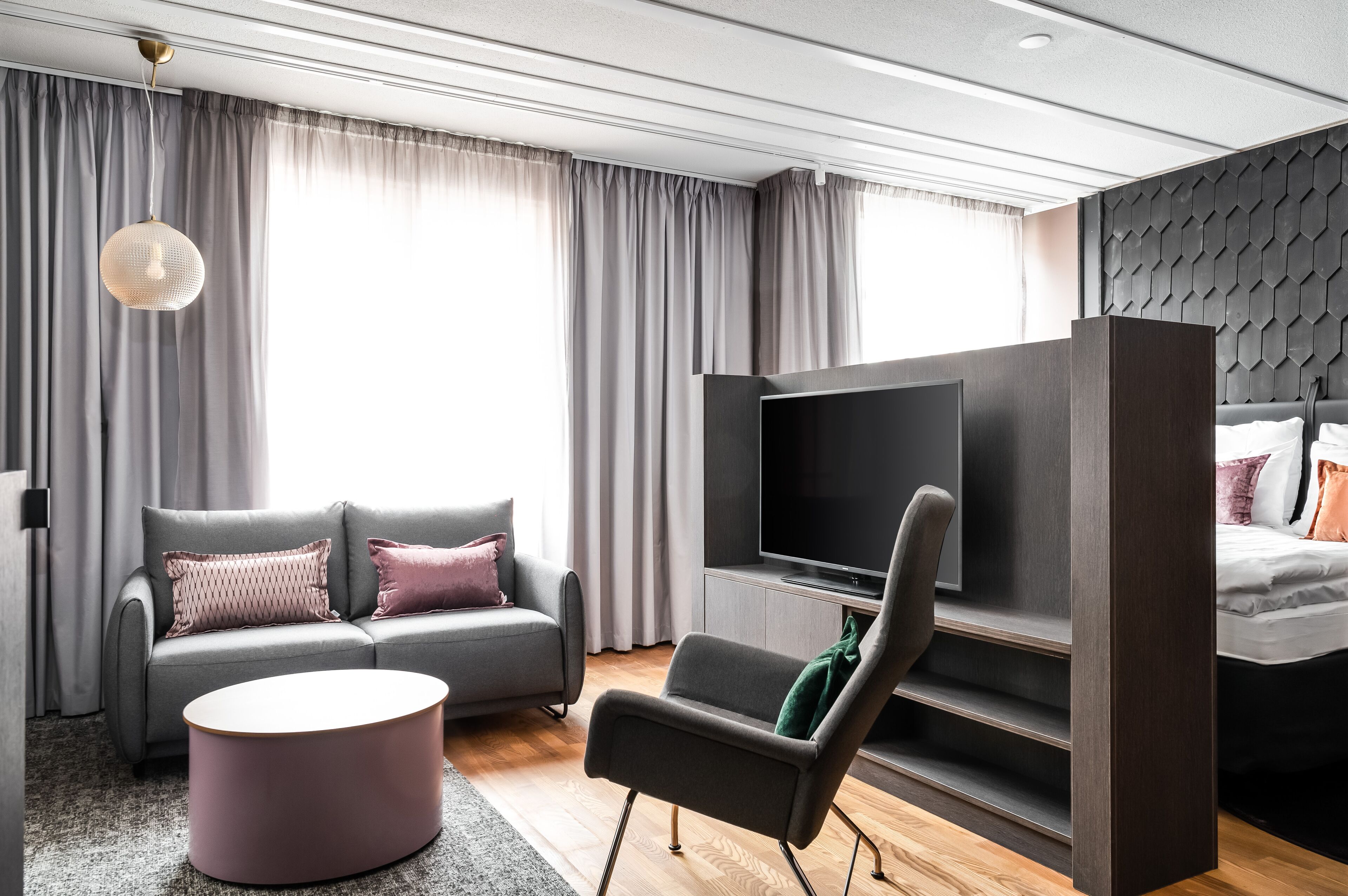 A modern hotel room with a grey sofa, a flat-screen TV, a small round table, and a bed with a dark headboard.