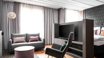 A modern hotel room with a grey sofa, a flat-screen TV, a small round table, and a bed with a dark headboard.