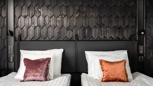 Ett hotellrum med två sängar, båda med vita sängkläder och en enkel, dekorativ prydnadskudde. Rummet har en mörk, strukturerad väggbeklädnad och en modern, minimalistisk inredning.