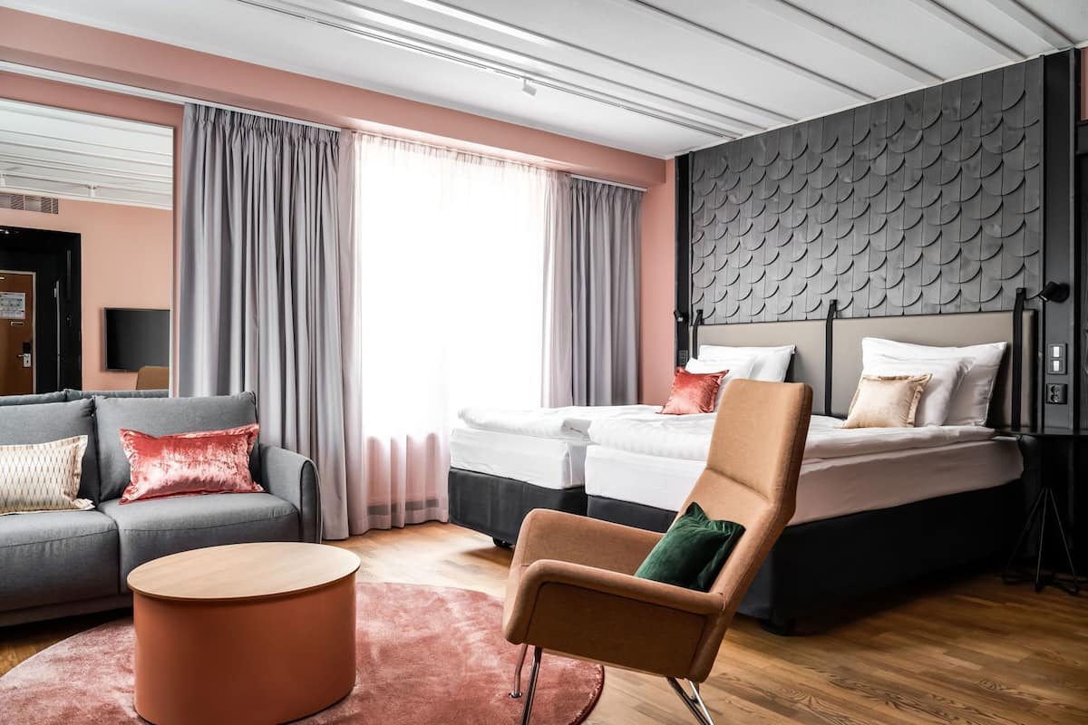 Une chambre d’hôtel moderne, équipée d’un canapé, de deux lits, d’une chaise, d’une table ronde et d’une grande fenêtre avec des rideaux.