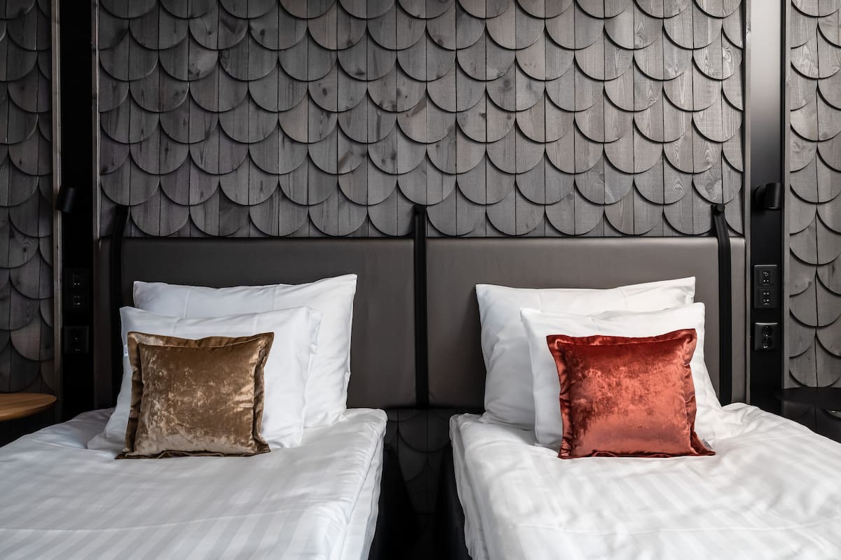 Une chambre d’hôtel avec un lit doté d’une tête de lit ornée d’un motif évoquant des écailles et de deux oreillers, l’un marron et l’autre rouge.