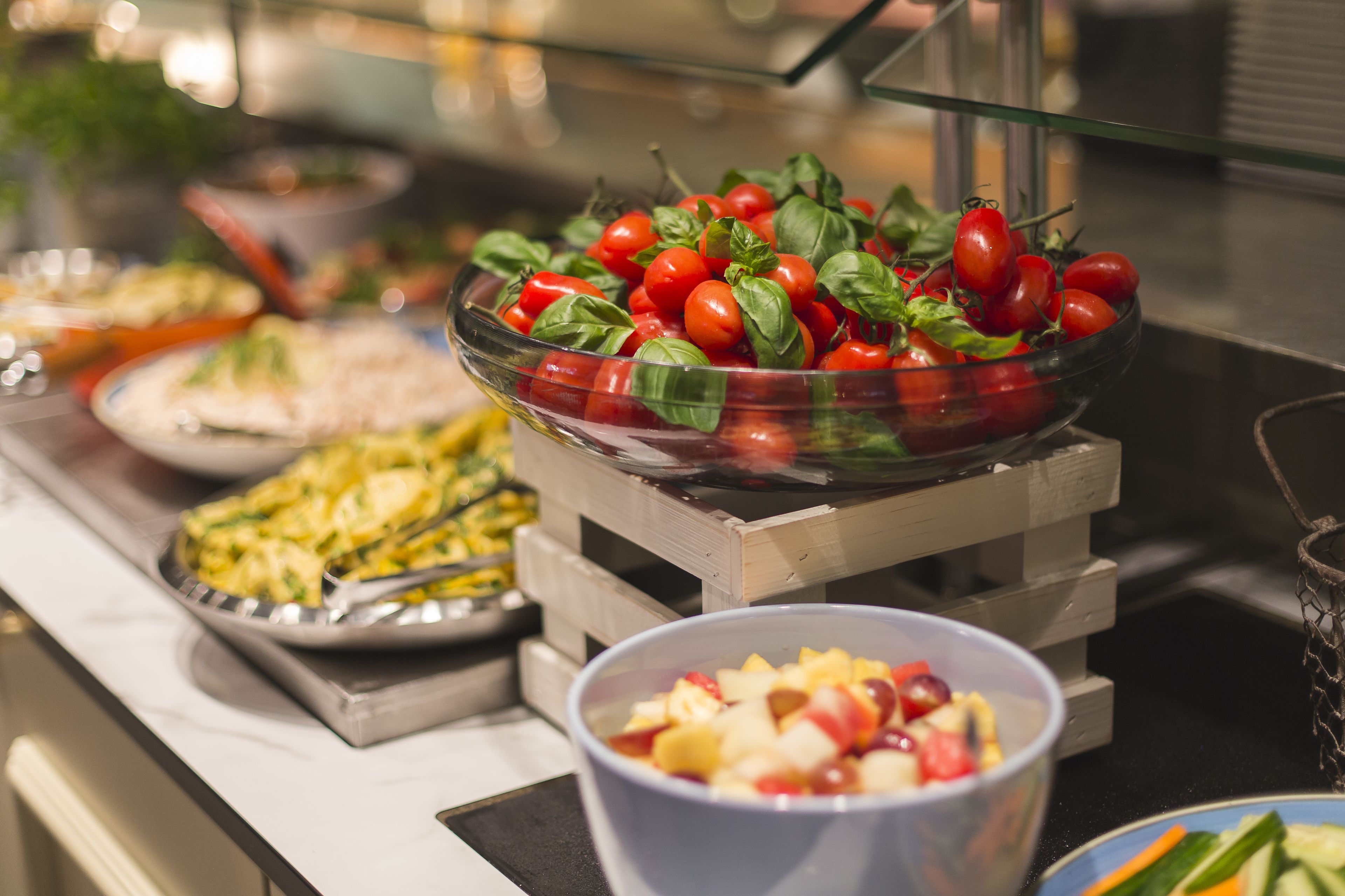 Un bufé con diversos platos, incluyendo un bol de tomates cherry y albahaca, un plato de pasta y un bol de ensalada de frutas.