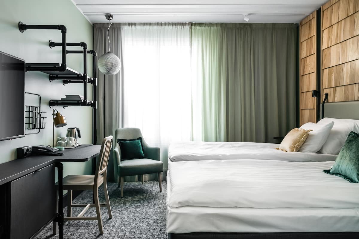 Une chambre d’hôtel équipée d’un bureau, de deux lits, d’une chaise, d’une télévision et d’une fenêtre avec des rideaux.