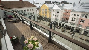 En balkong med trägolv, en stol och en blomsteruppsättning. I bakgrunden syns färgglada byggnader och en gata med människor och cyklar.