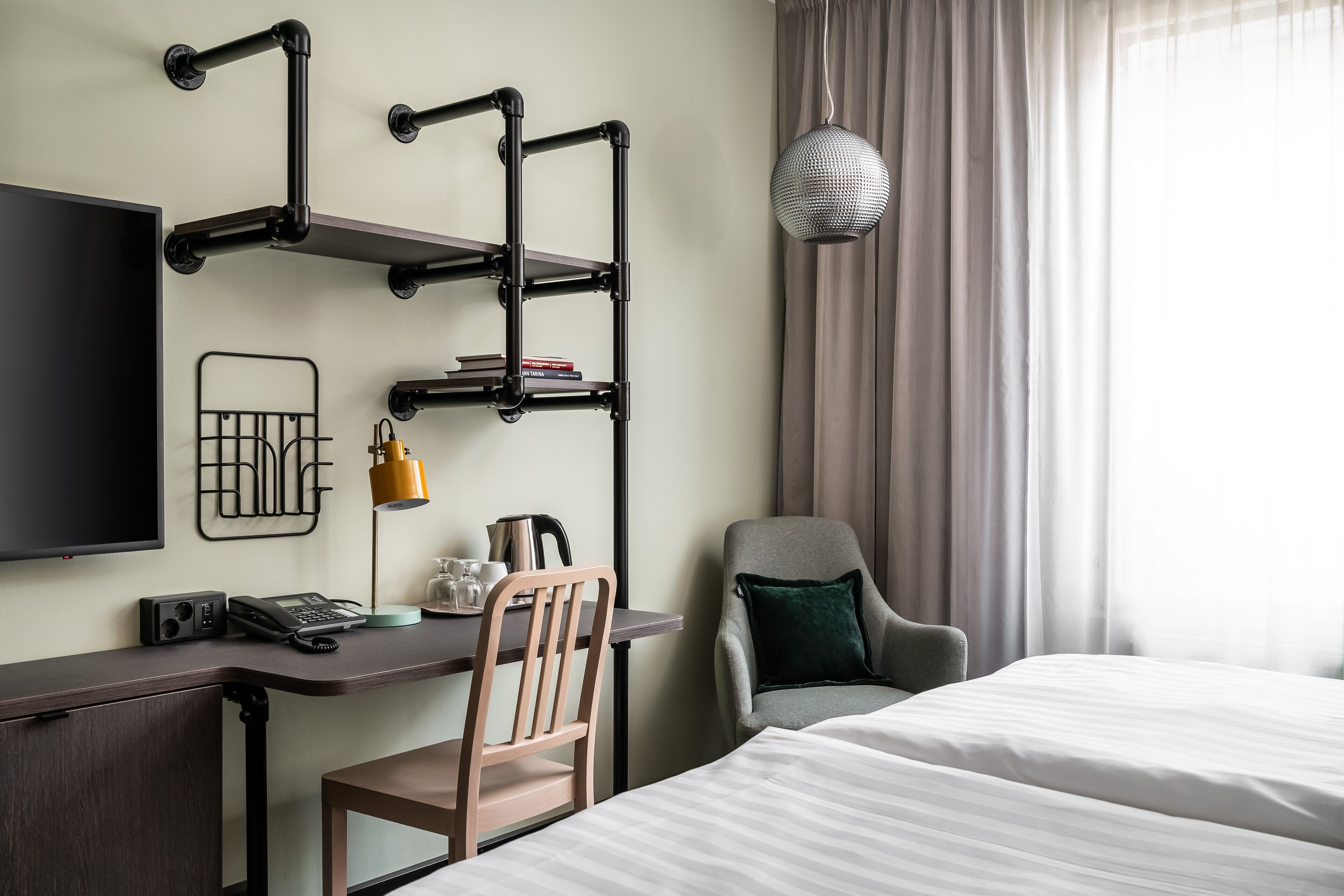 Une chambre d’hôtel comprenant un lit, un bureau avec une chaise, une télévision, une étagère avec des livres, une lampe et une fenêtre avec des rideaux.