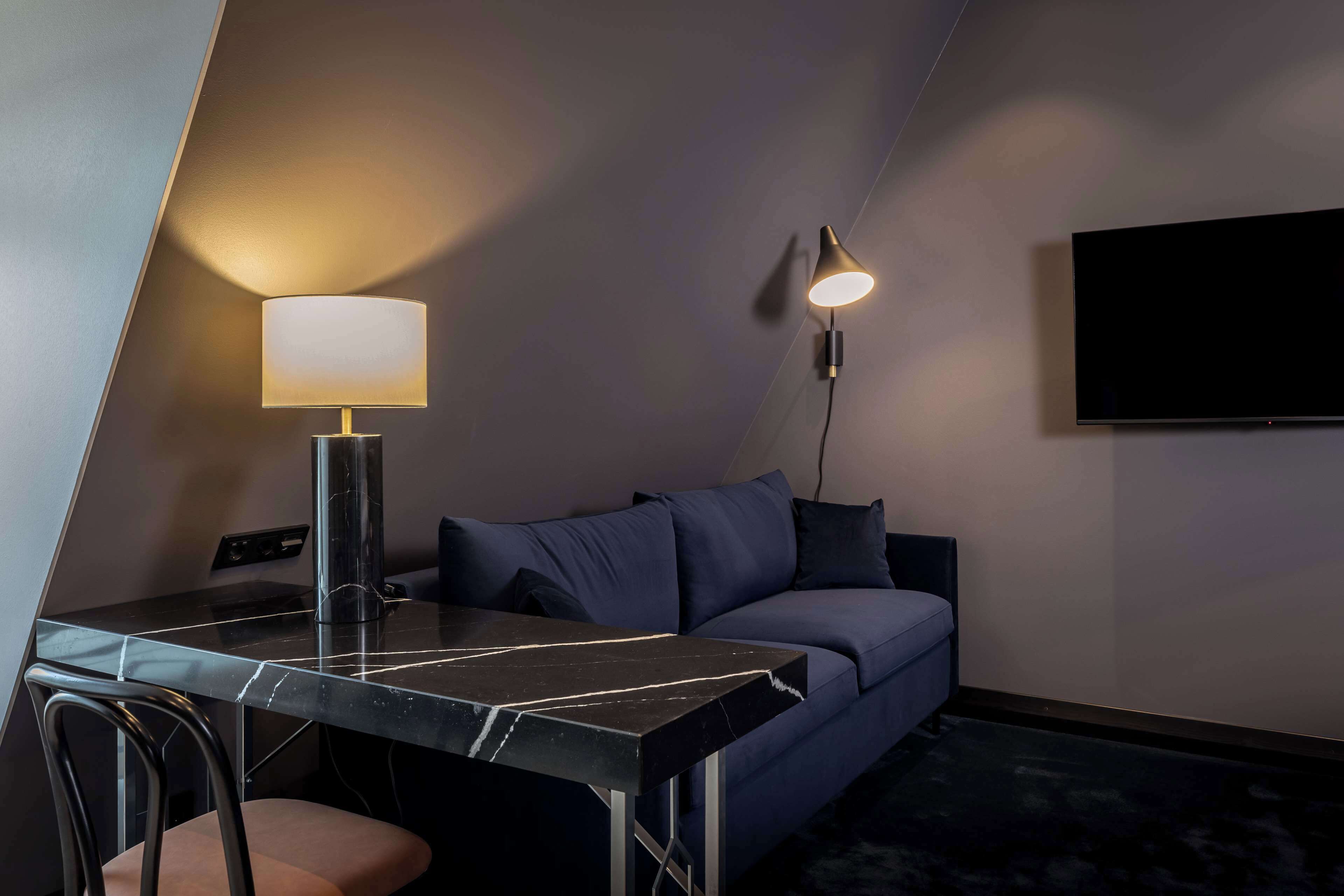 A modern hotel room with a dark blue sofa, a marble table, a lamp, and a flat-screen TV mounted on the wall.