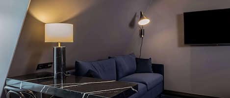 A modern hotel room with a dark blue sofa, a marble table, a lamp, and a flat-screen TV mounted on the wall.