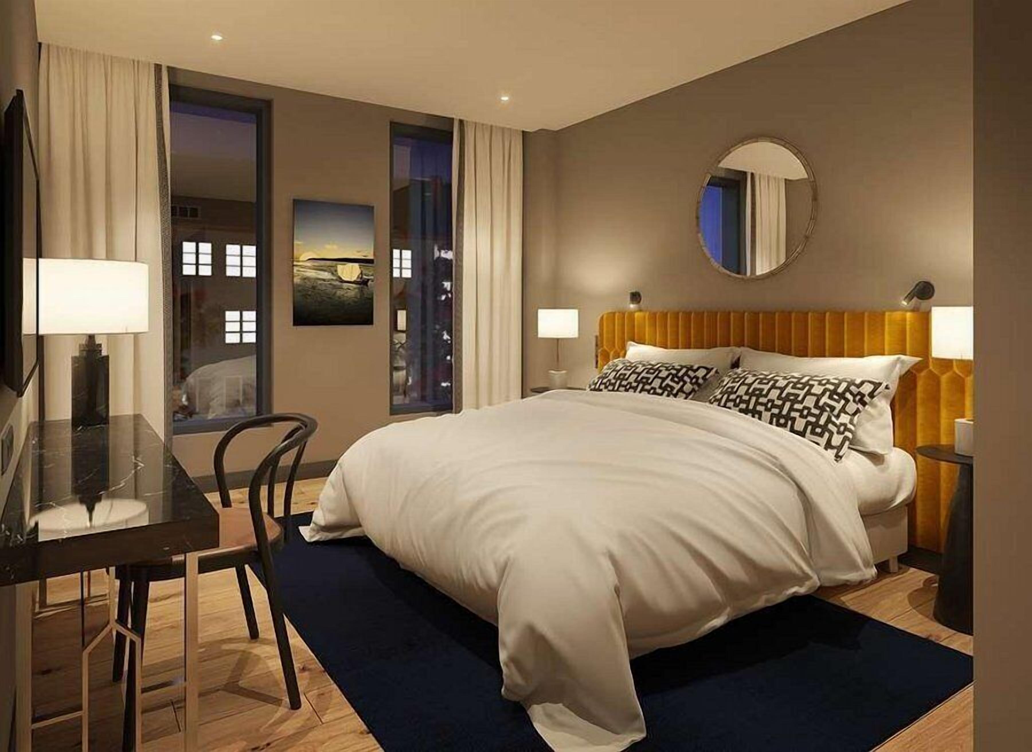 A modern hotel room with a large bed, a desk, and a chair. There are two bedside tables with lamps, a round mirror on the wall, and a window with curtains.