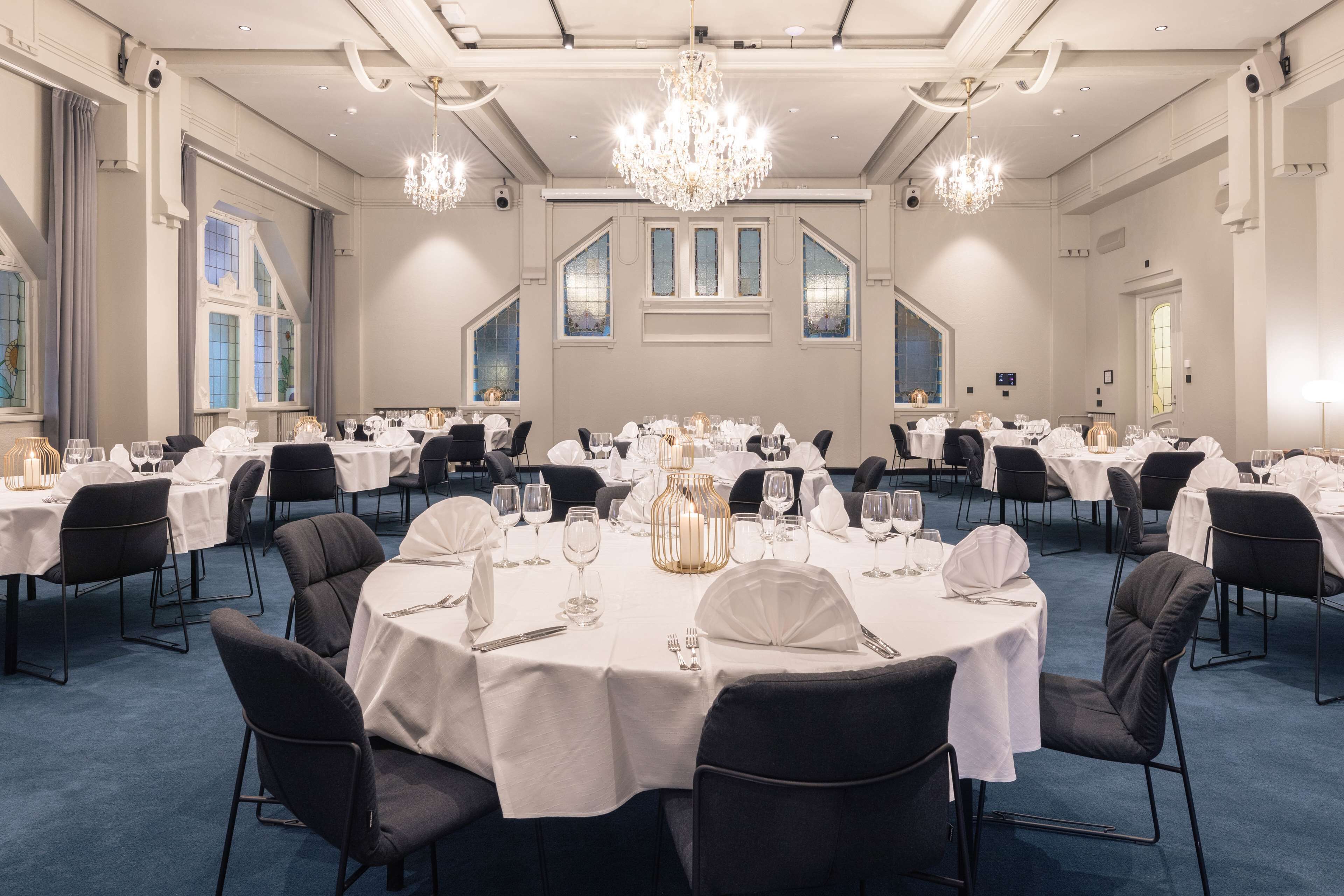 Un amplio salón de banquetes con mesas redondas dispuestas para comer, candelabros y grandes ventanales.