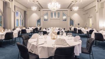 Un amplio salón de banquetes con mesas redondas dispuestas para comer, candelabros y grandes ventanales.