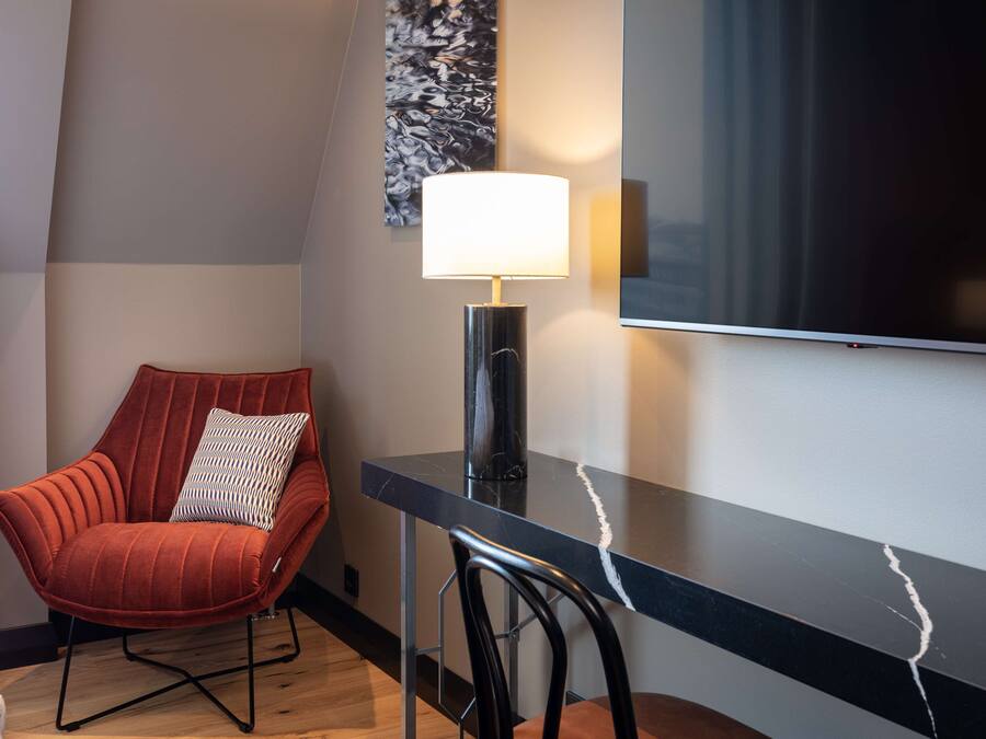 Una habitación de hotel moderna con una butaca tapizada de rojo, un escritorio negro con una lámpara y un televisor de pantalla plana grande montado en la pared.