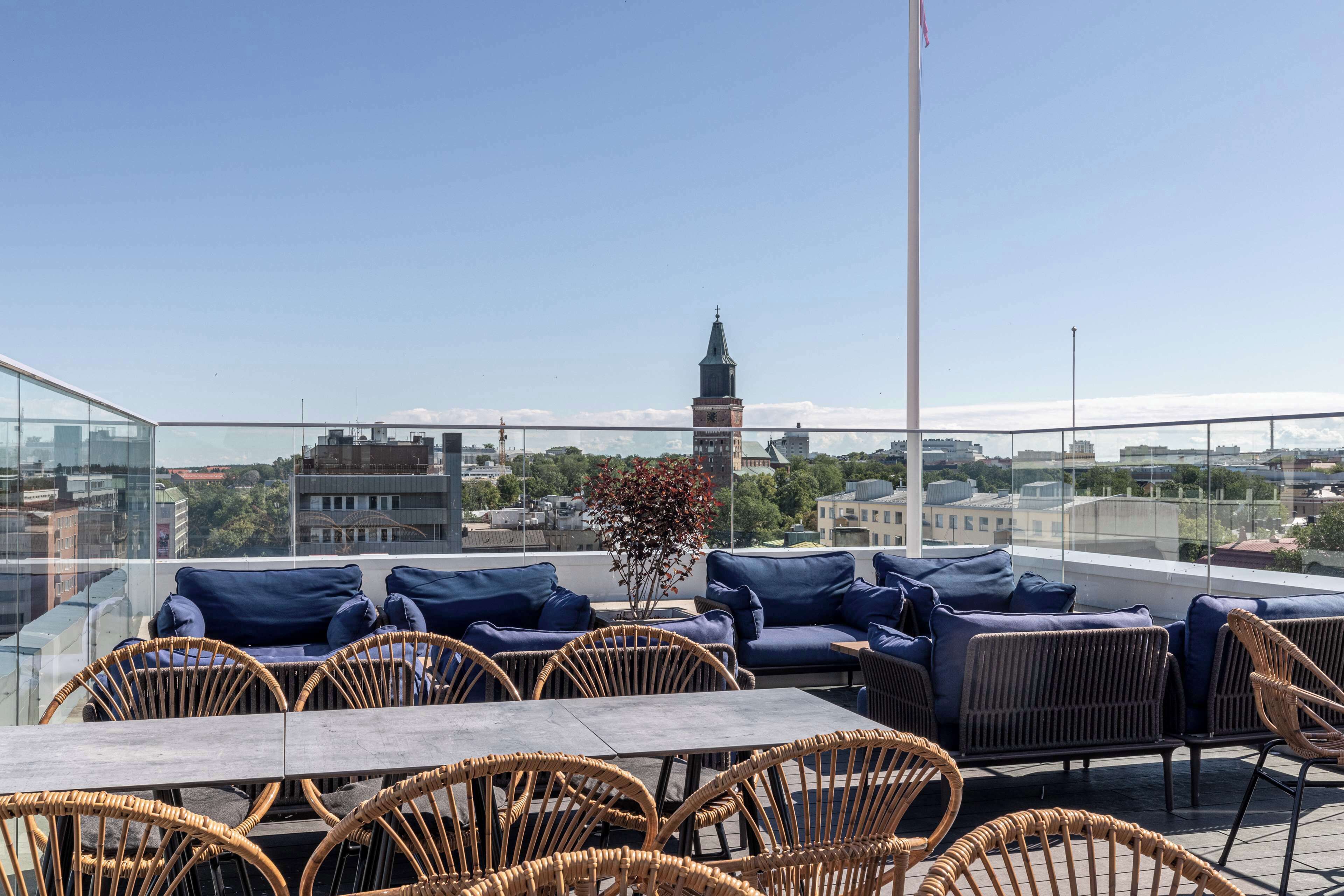 A rooftop terrace with wicker chairs and blue cushions, overlooking a cityscape with a prominent tower.