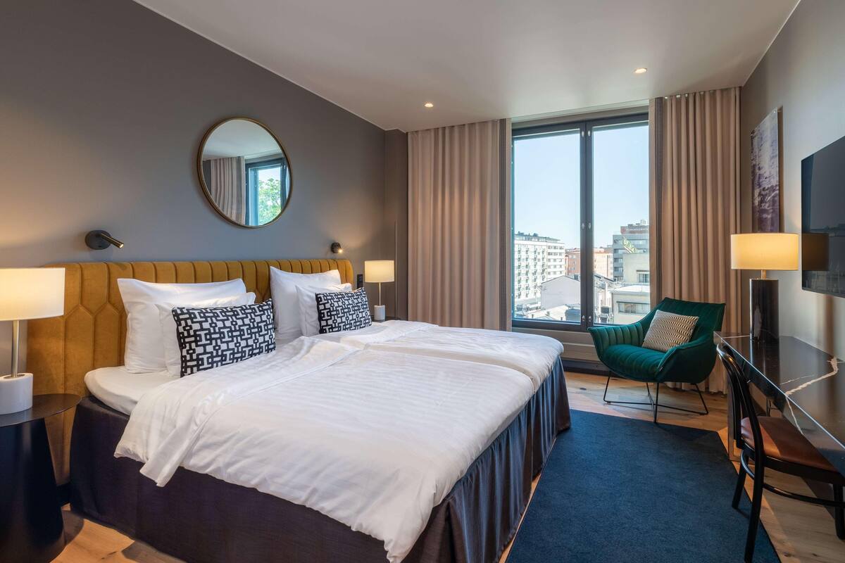 A hotel room with a large bed, two bedside tables, a green armchair, a wooden bench, a large window with curtains, and a flat-screen TV.