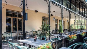 Un café al aire libre con mesas y sillas, plantas en macetas y un balcón con barandillas.