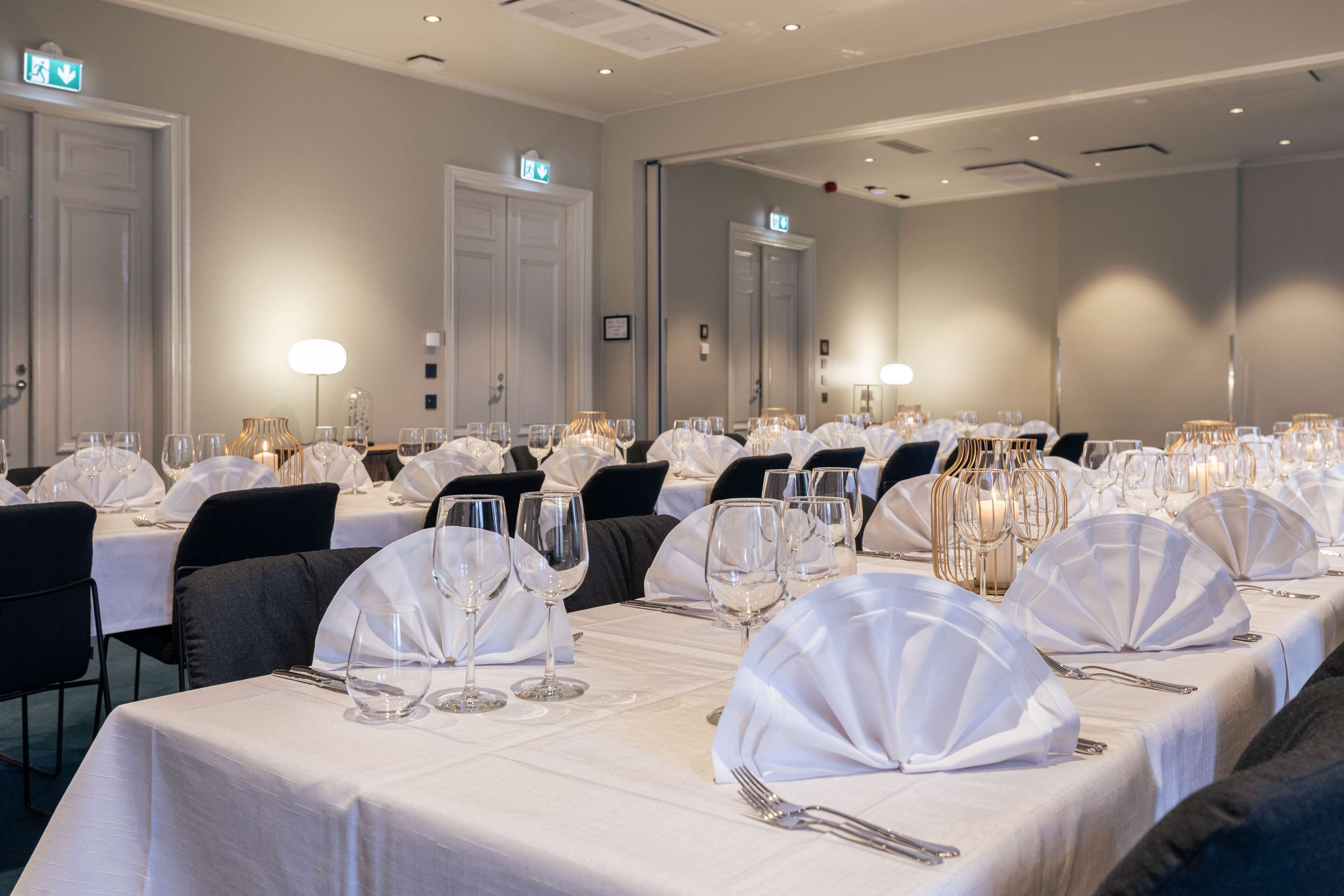 Una sala de banquetes amb diverses taules preparades per a dinar, cadascuna amb teles de taula blanques, vetrería i espelmes daurades.