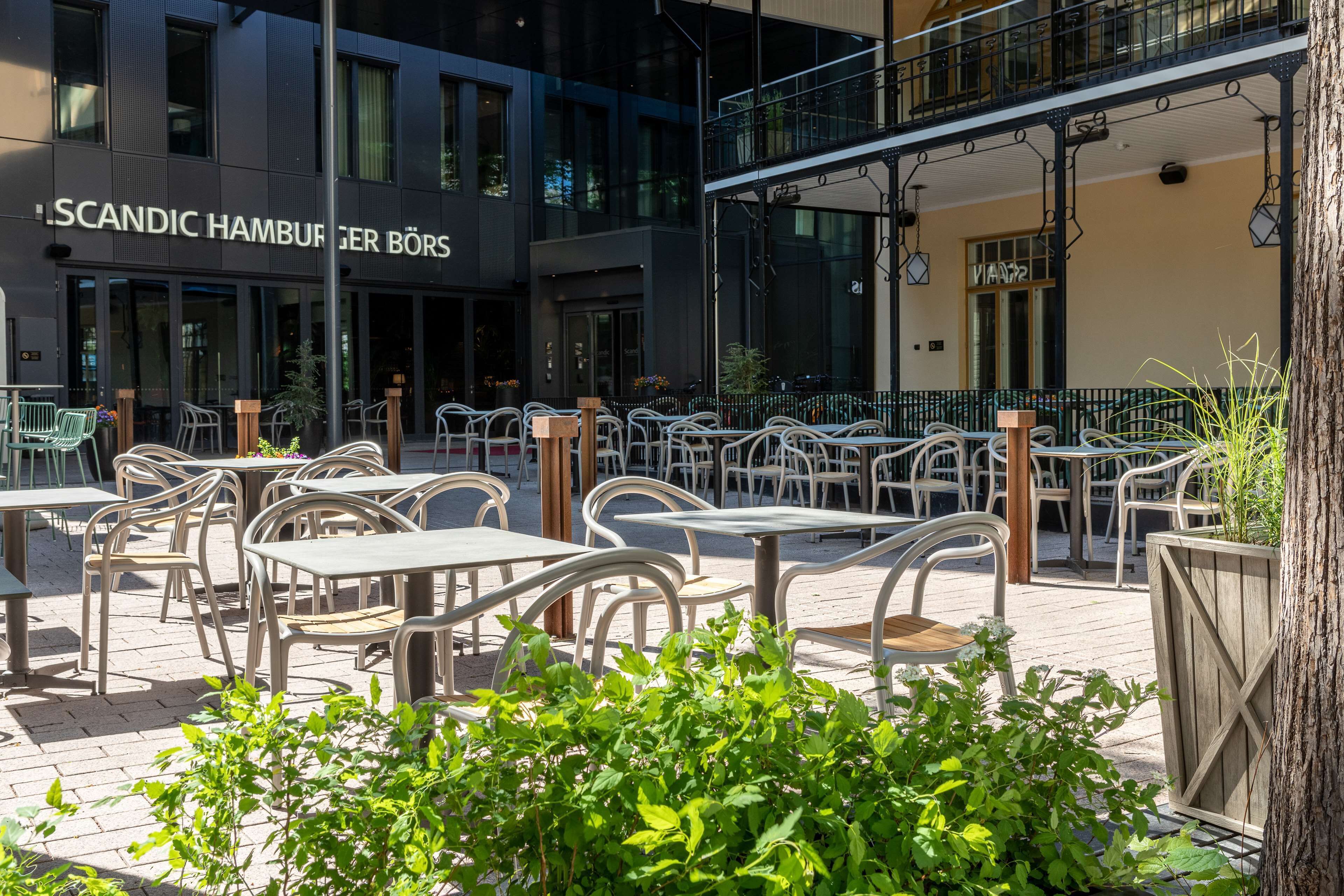 Un espace extérieur aménagé avec des tables et des chaises, un bâtiment sur lequel on peut lire l’inscription « SCANDIC HAMBURGER BÖRS », et un arbre au premier plan.