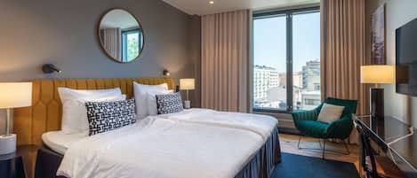 Une chambre d’hôtel avec un grand lit, deux tables de chevet, un fauteuil vert, une chaise en bois et une grande fenêtre avec des rideaux.