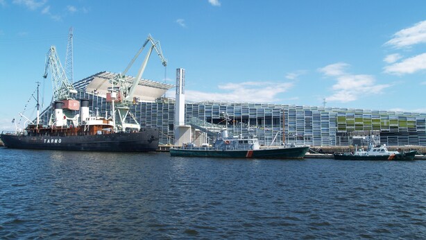 Un gran buque de carga atracado en un puerto con grúas y un moderno edificio industrial al fondo.