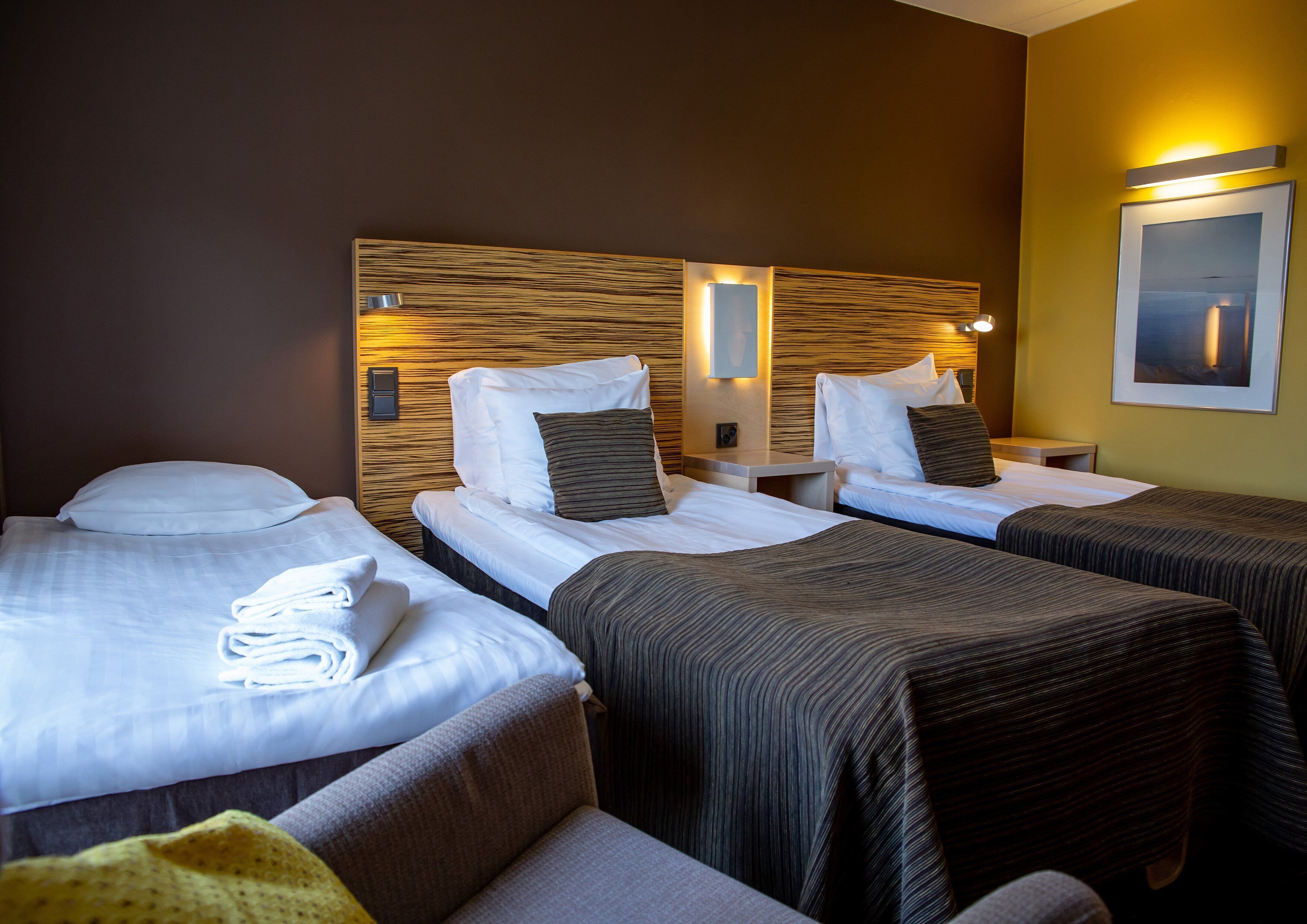 Habitación de hotel con dos camas, cada una con sábanas y almohadas blancas, cabeceros de madera, mesitas de noche y un cuadro en la pared.