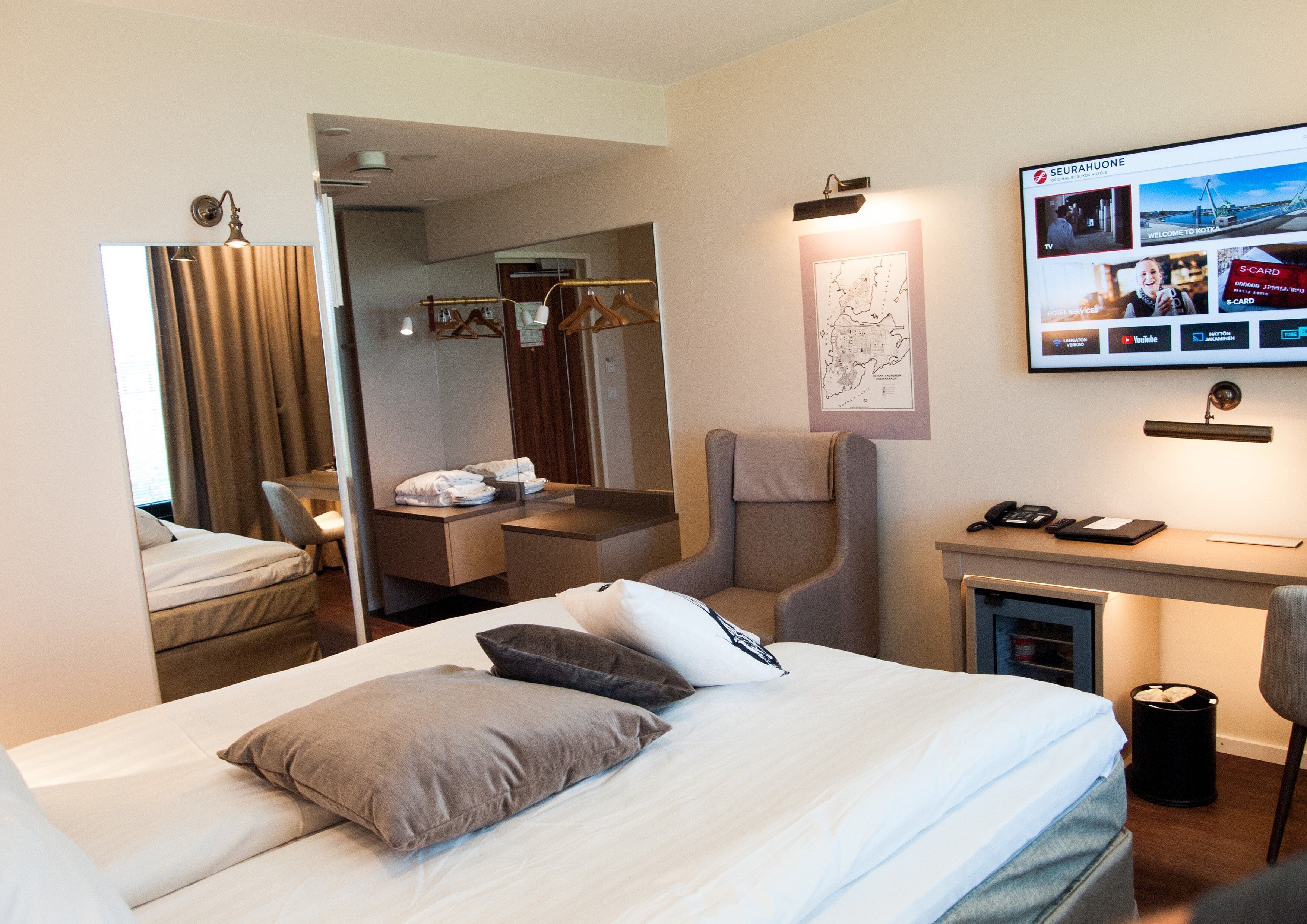A hotel room with a bed, a desk, a chair, a mirror, and a TV displaying a website.
