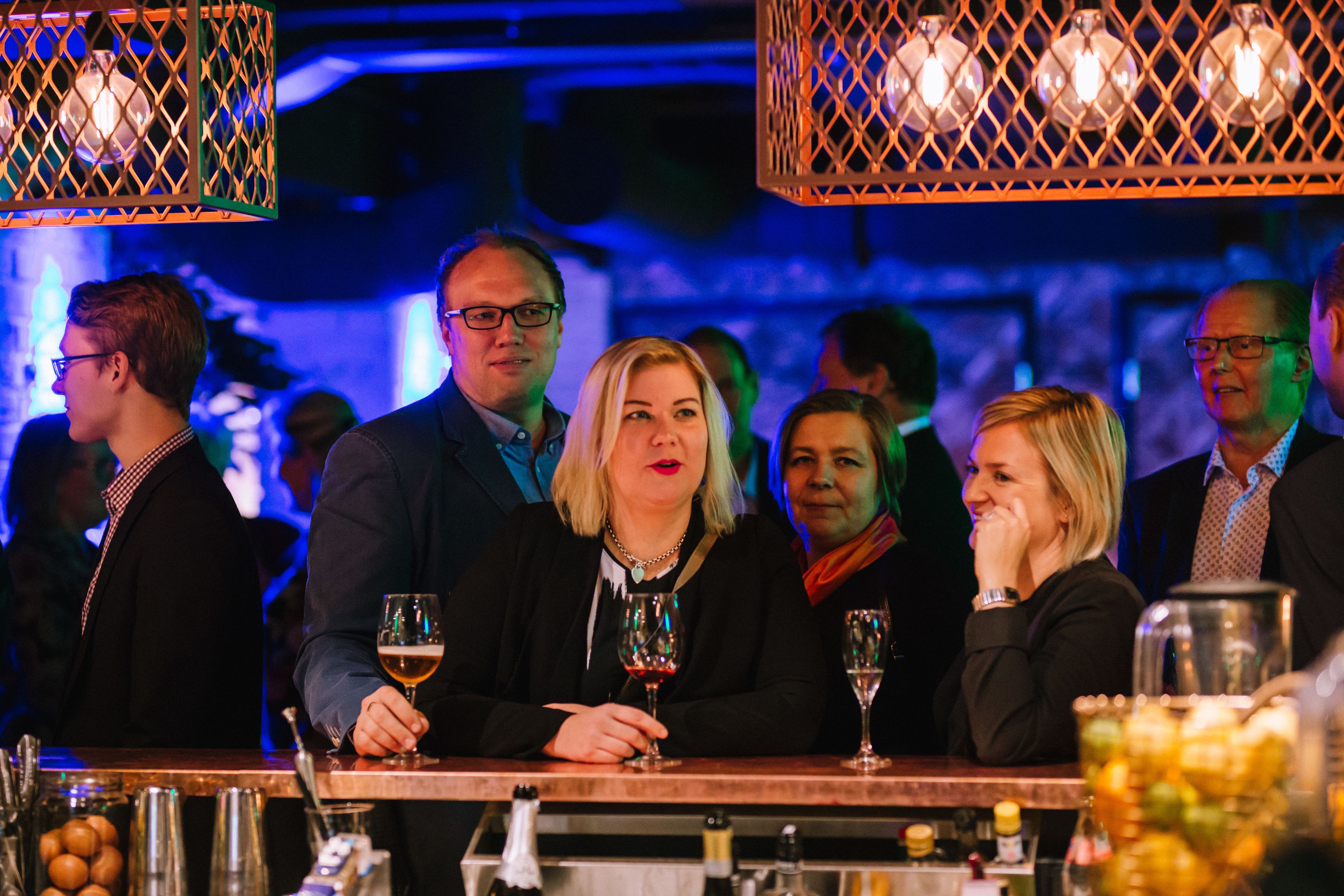 A bar setting with people seated at tables, glasses of wine, and a bar counter with bottles and glasses.