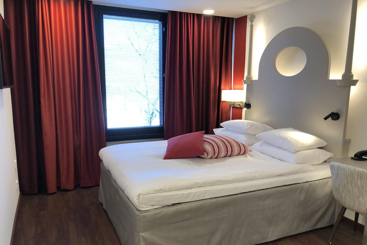 Una habitación de hotel con una cama grande, cortinas rojas, una ventana con persianas, una lámpara de pared y un elemento arquitectónico circular.
