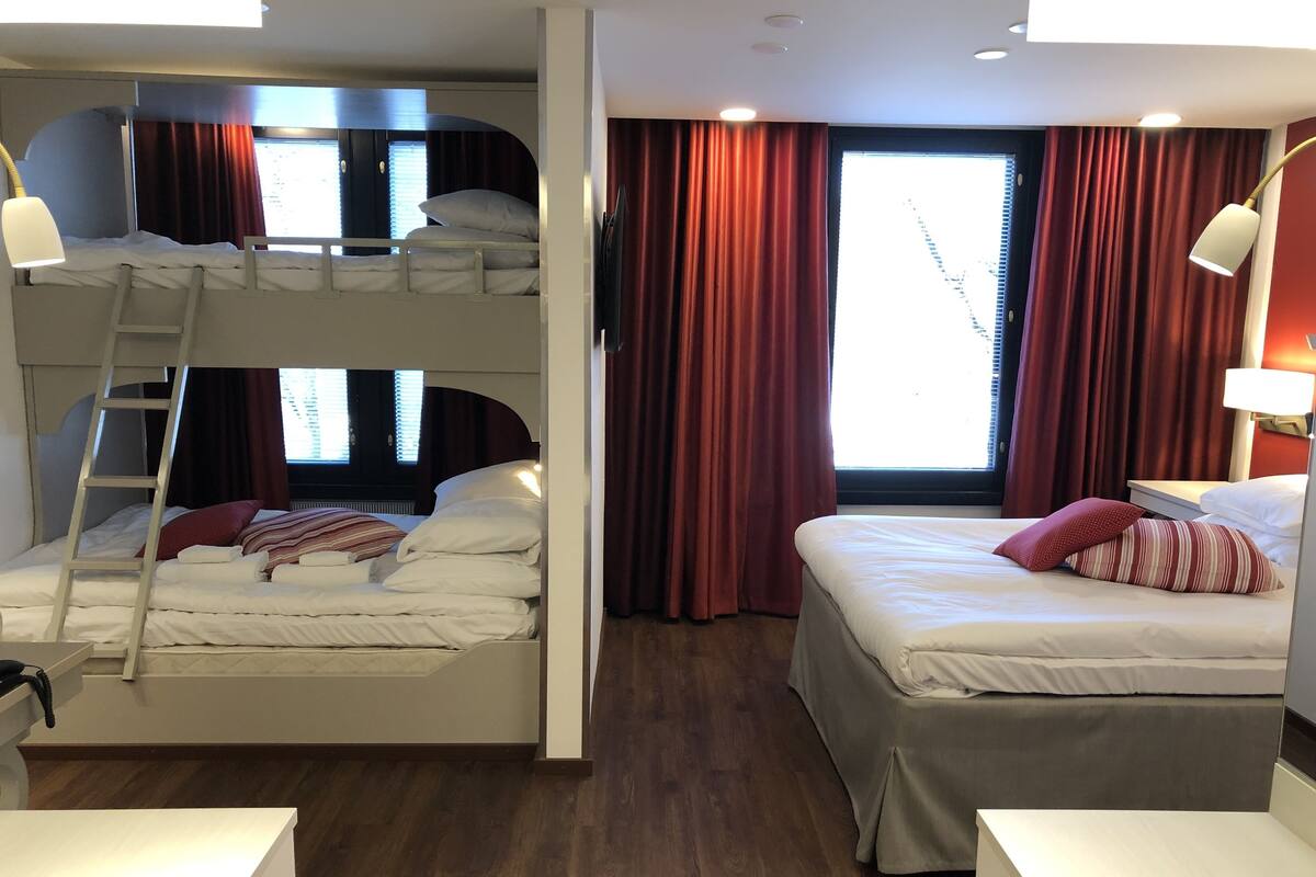 Una habitación con dos camas, una litera y otra individual, ambas con ropa de cama y almohadas blancas. Incluye una escalera para la litera, un escritorio con silla y una ventana con cortinas.