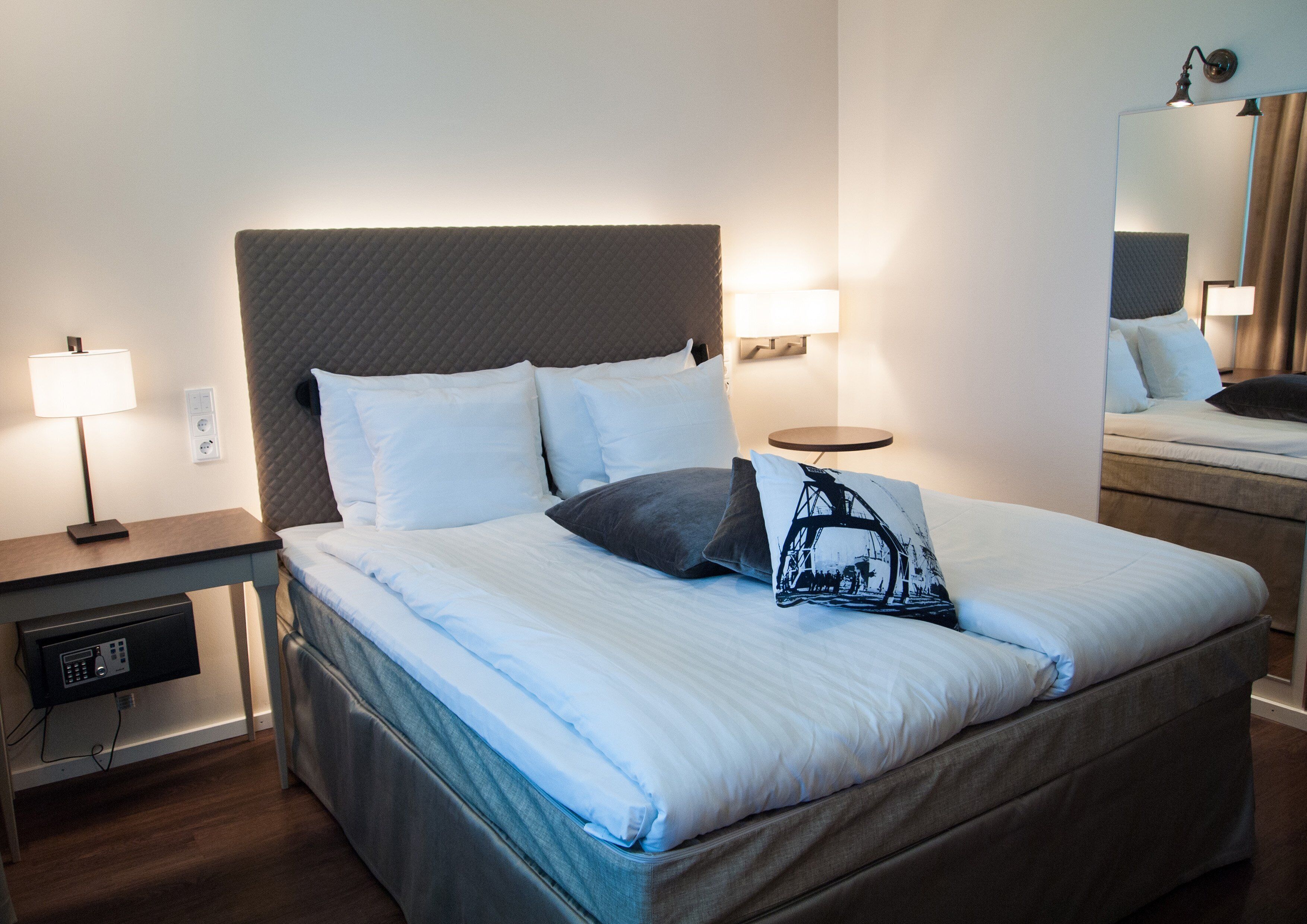 Una cama bien hecha con sábanas blancas y un cojín blanco y negro, una mesita de noche con una lámpara y un televisor pequeño junto a la cama.