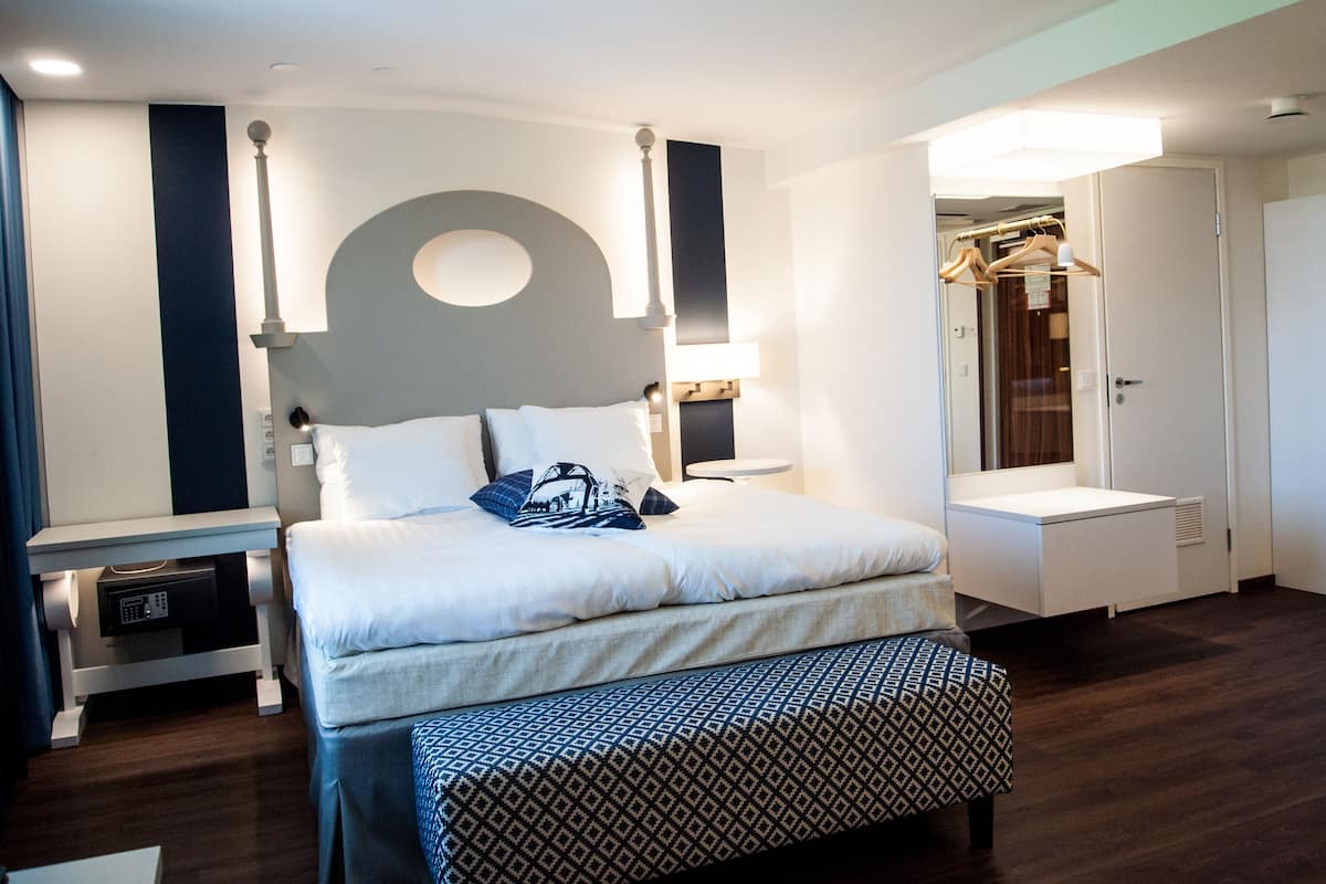 Una habitación de hotel moderna con cama, mesitas de noche, un banco, un espejo y un armario.