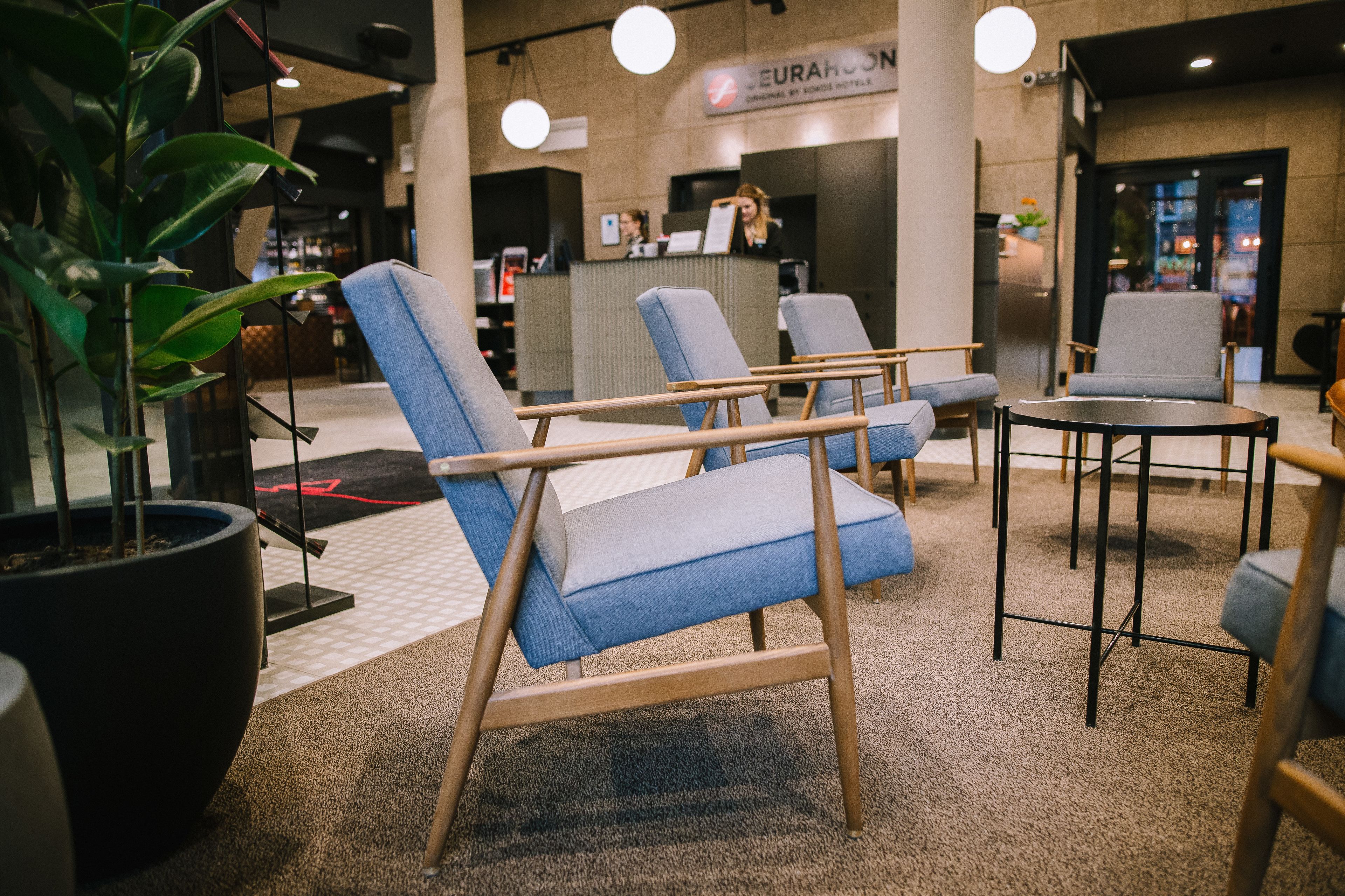 Een zithoek met blauwe fauteuils en een ronde tafel, een receptiebalie en een potplant.