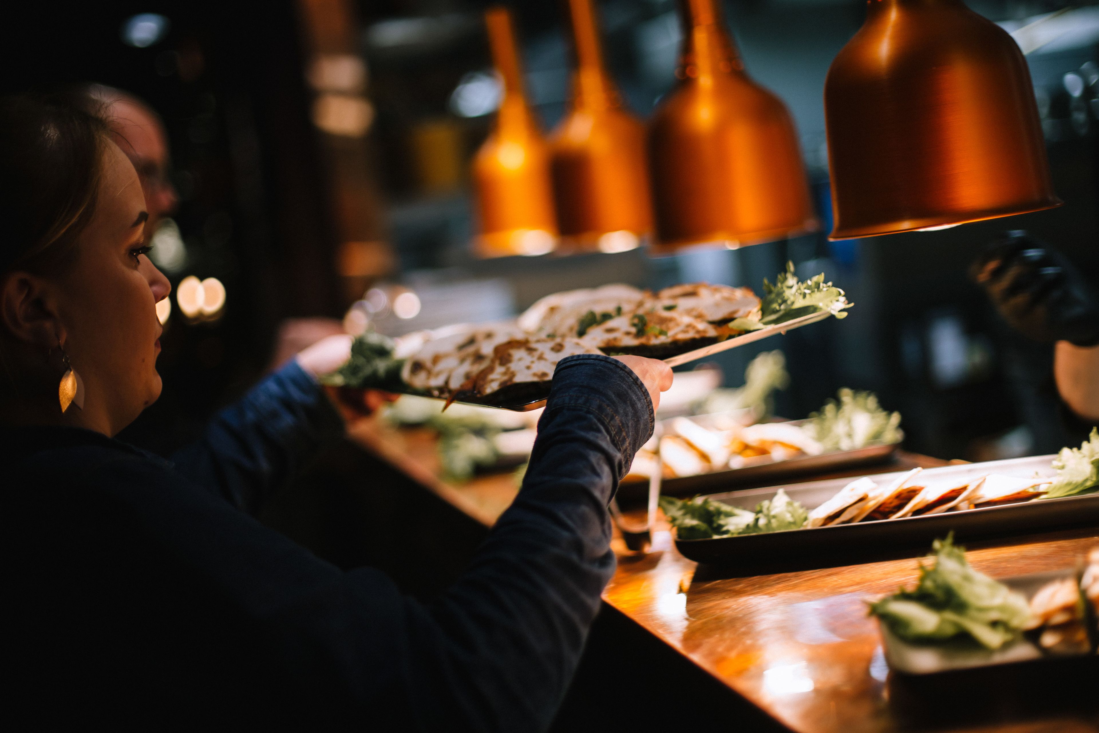 Une personne servant des plats dans un restaurant doté d’un comptoir en bois et de luminaires suspendus en cuivre.