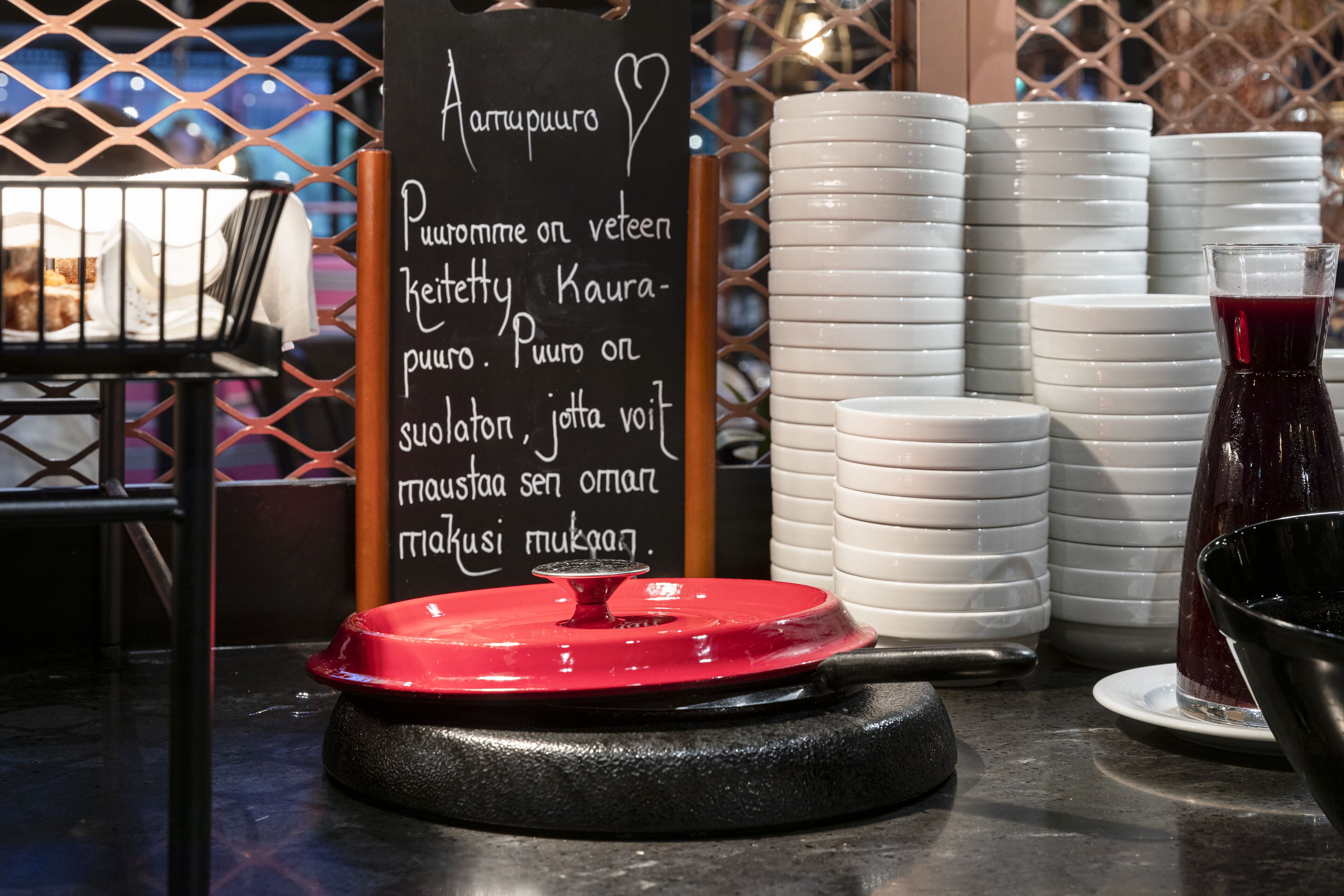 Montaje de bufé con un cartel que enumera los alimentos, platos blancos apilados y un recipiente de comida con tapa roja.