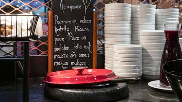 Montaje de bufé con un cartel que enumera los alimentos, platos blancos apilados y un recipiente de comida con tapa roja.