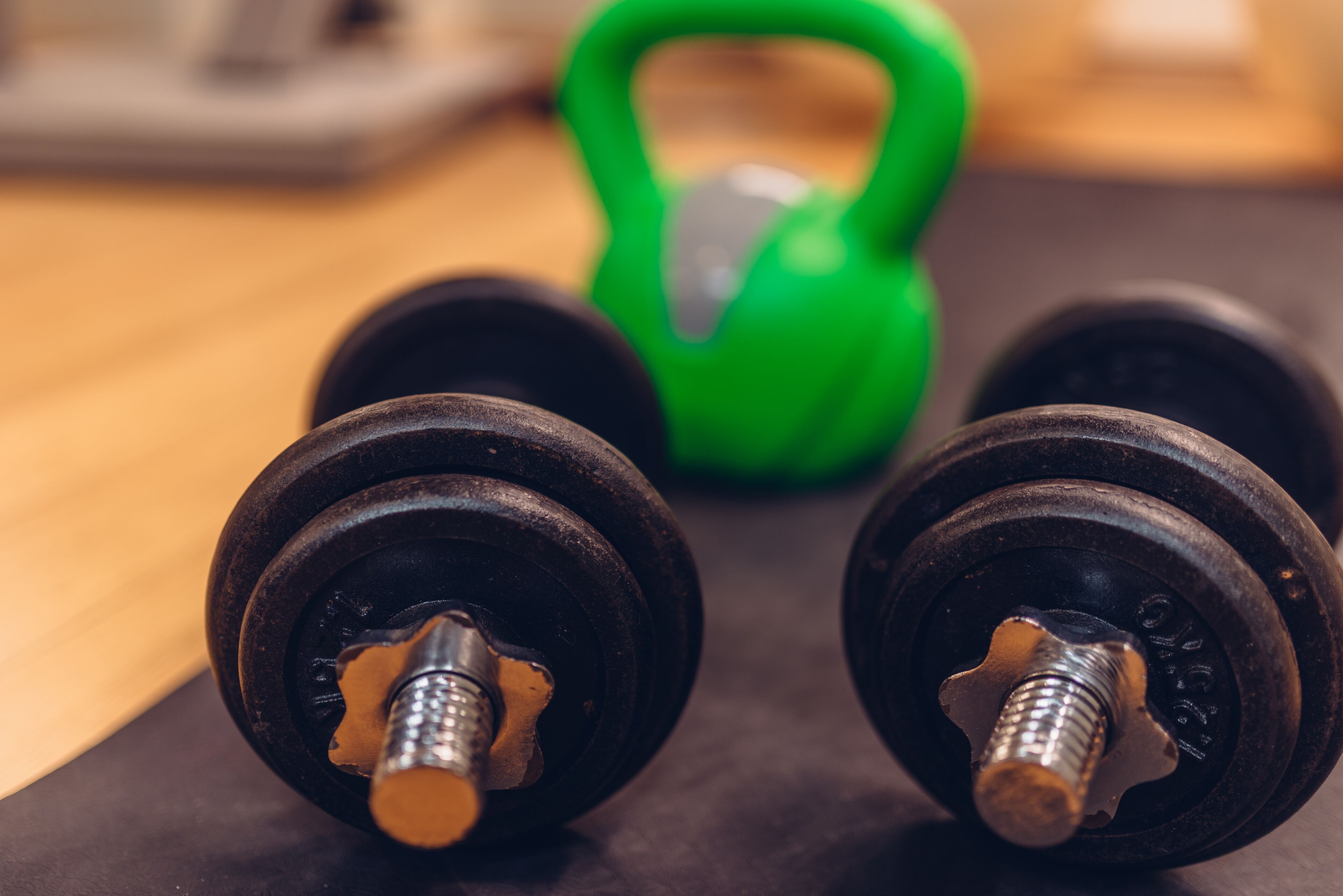 Eine Nahaufnahme von zwei schwarzen Kurzhanteln mit silbernen Gewichten auf einer schwarzen Matte, im Hintergrund eine grüne Kettlebell.