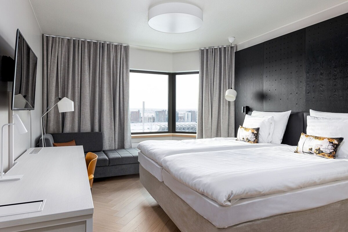 A modern hotel room with a large bed, a desk, a chair, and a view of the city.