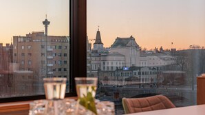 Una mesa con vasos y un florero, una ventana con vista a edificios y un puente, y una silla.