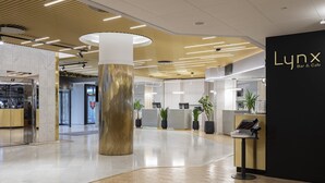 Um interior moderno com uma área de recepção, uma grande coluna de madeira e uma parede preta com a identificação 'Lynx Bar & Cafe'.