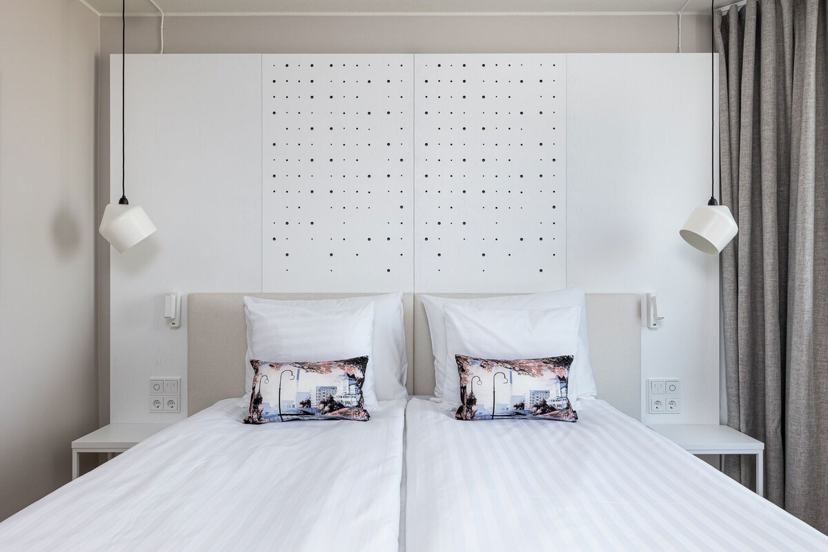 A double bed with white bedding and two decorative pillows, a white headboard with a dot pattern, bedside tables with lamps, and a grey curtain.